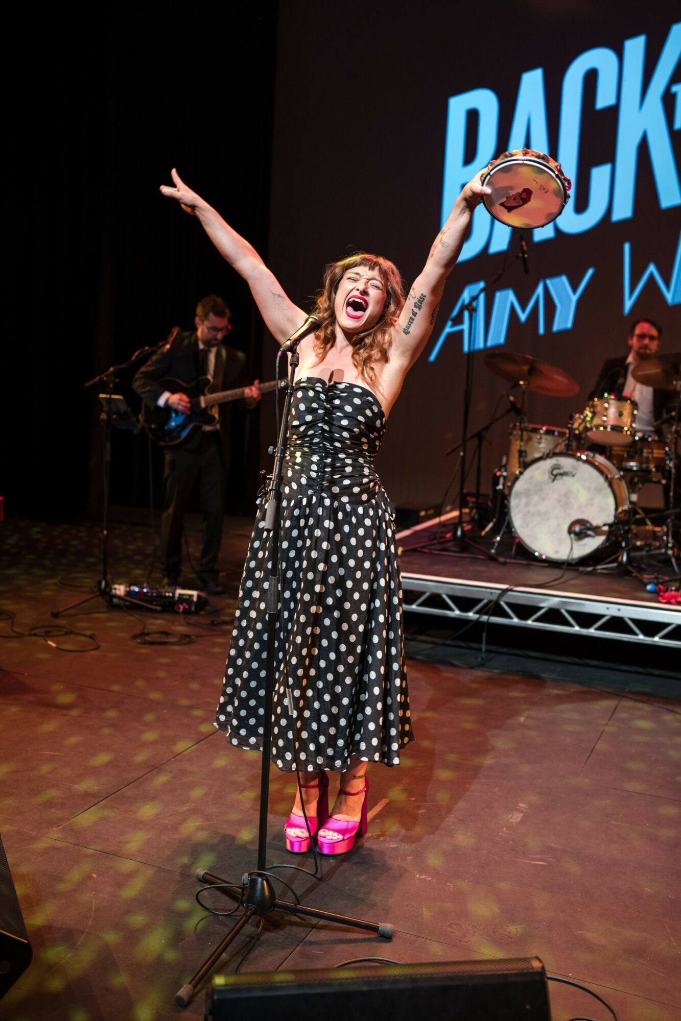 Performer with arms raised, holding a hat, singing on stage with band and large screen behind her.