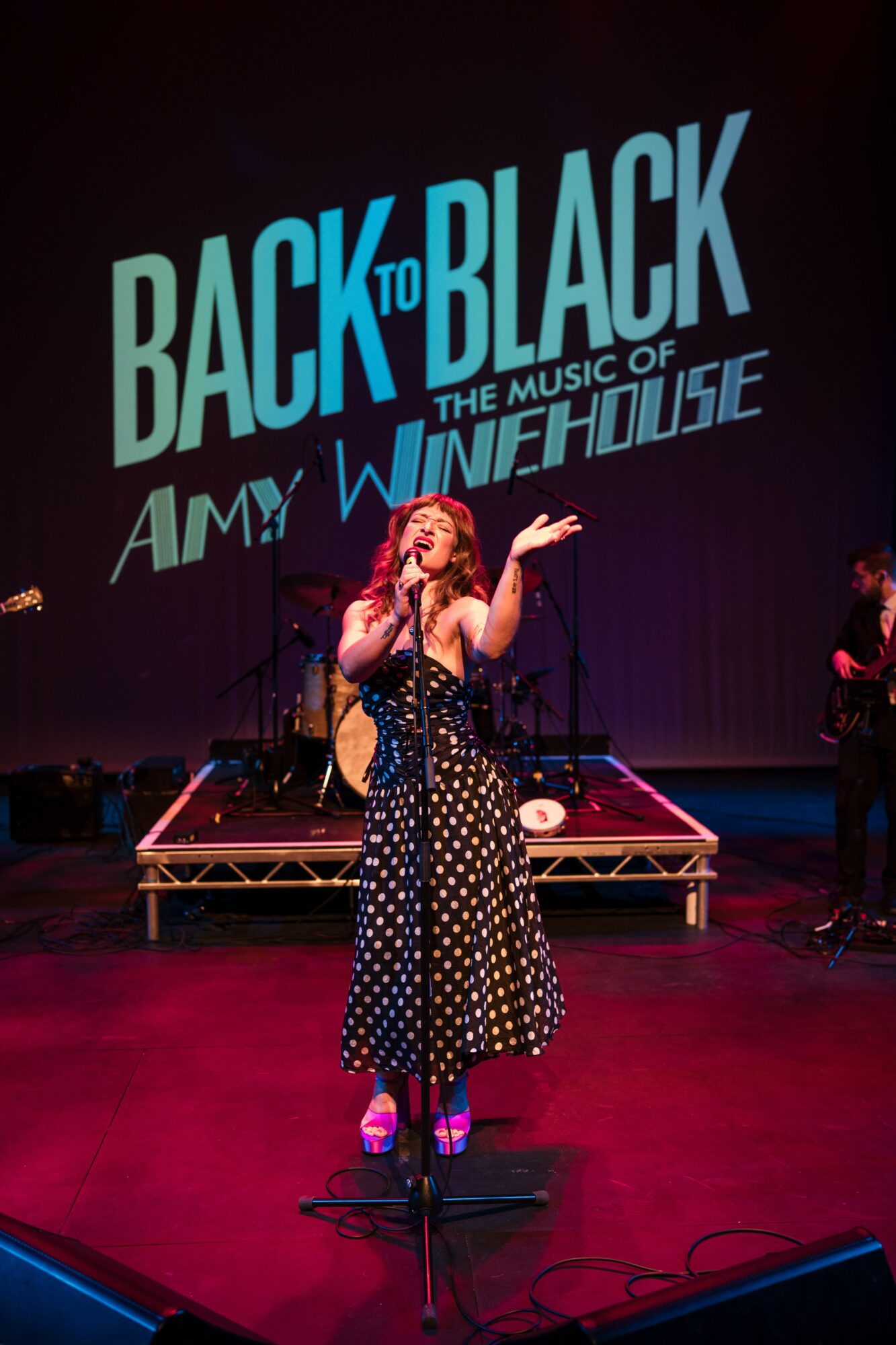 Performer singing on stage with a large screen behind displaying text about Amy Winehouse musical.