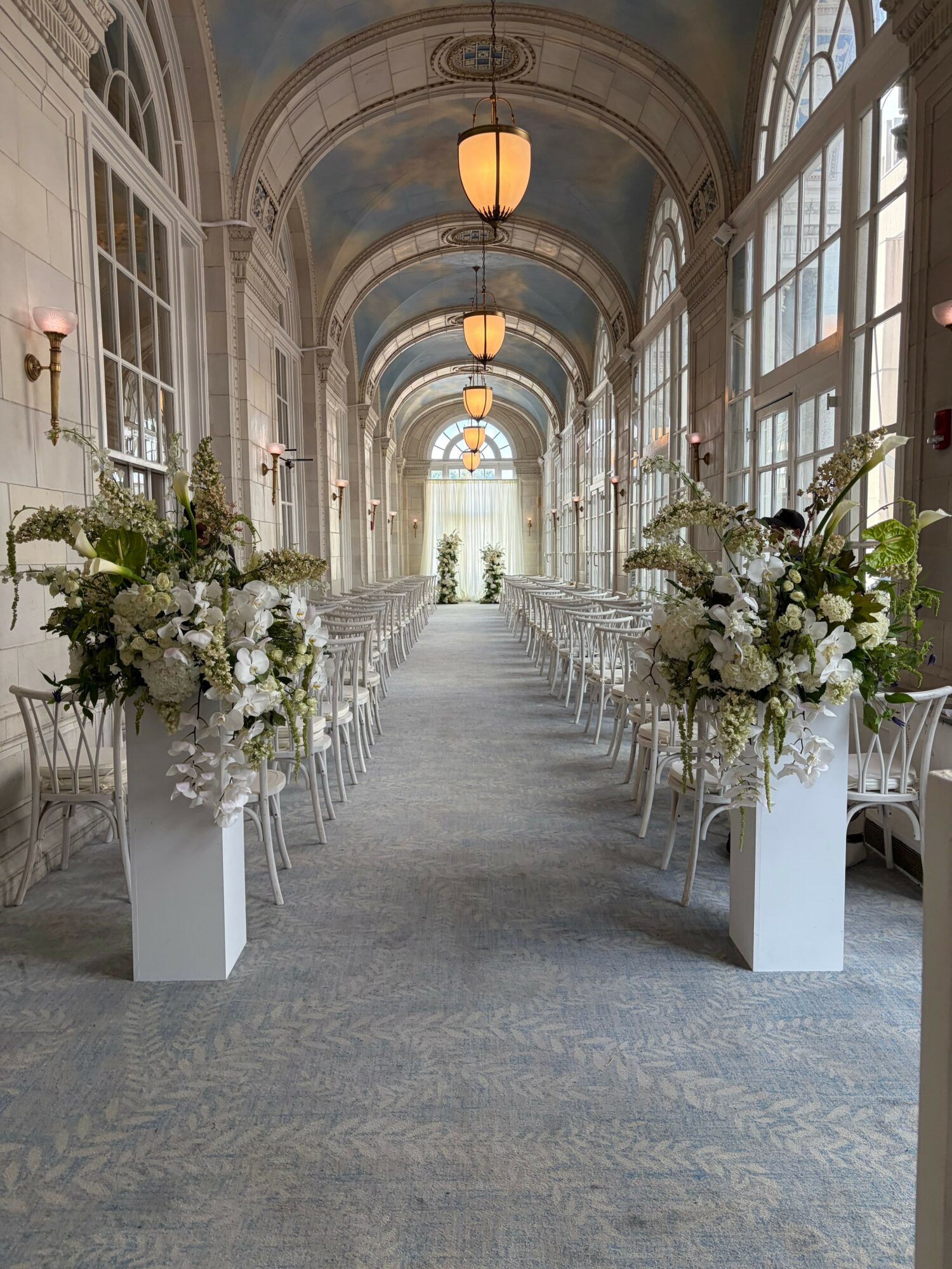 Elegant event space with floral arrangements on tall white stands, large windows, and a high arched ceiling.