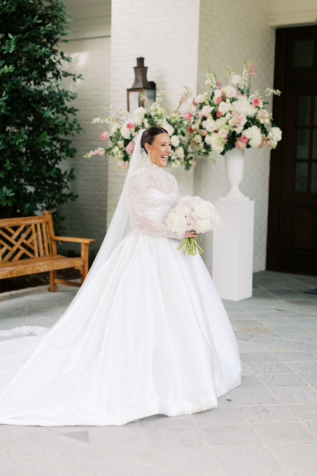 Bride in a white wedding dress holding a bouquet, standing outdoors near floral arrangements and a bench.