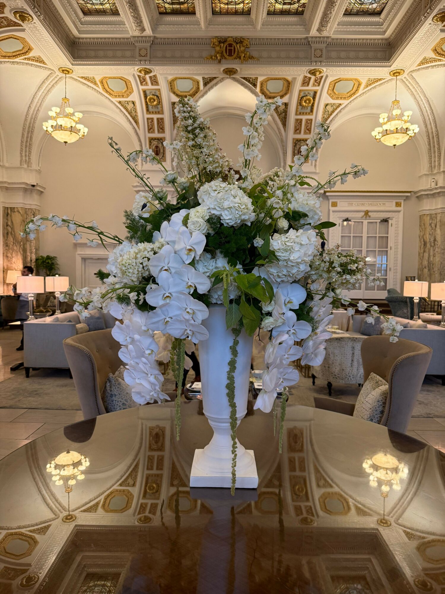 Elegant room with ornate ceiling, large floral arrangement with white and green flowers, and a table with a cake.