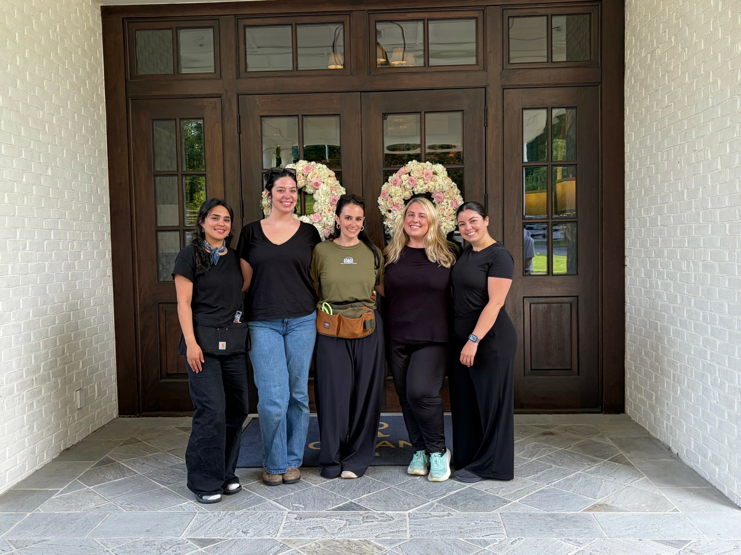Five women standing in front of a large wooden door with floral arrangements, smiling, indoors, tiled floor.