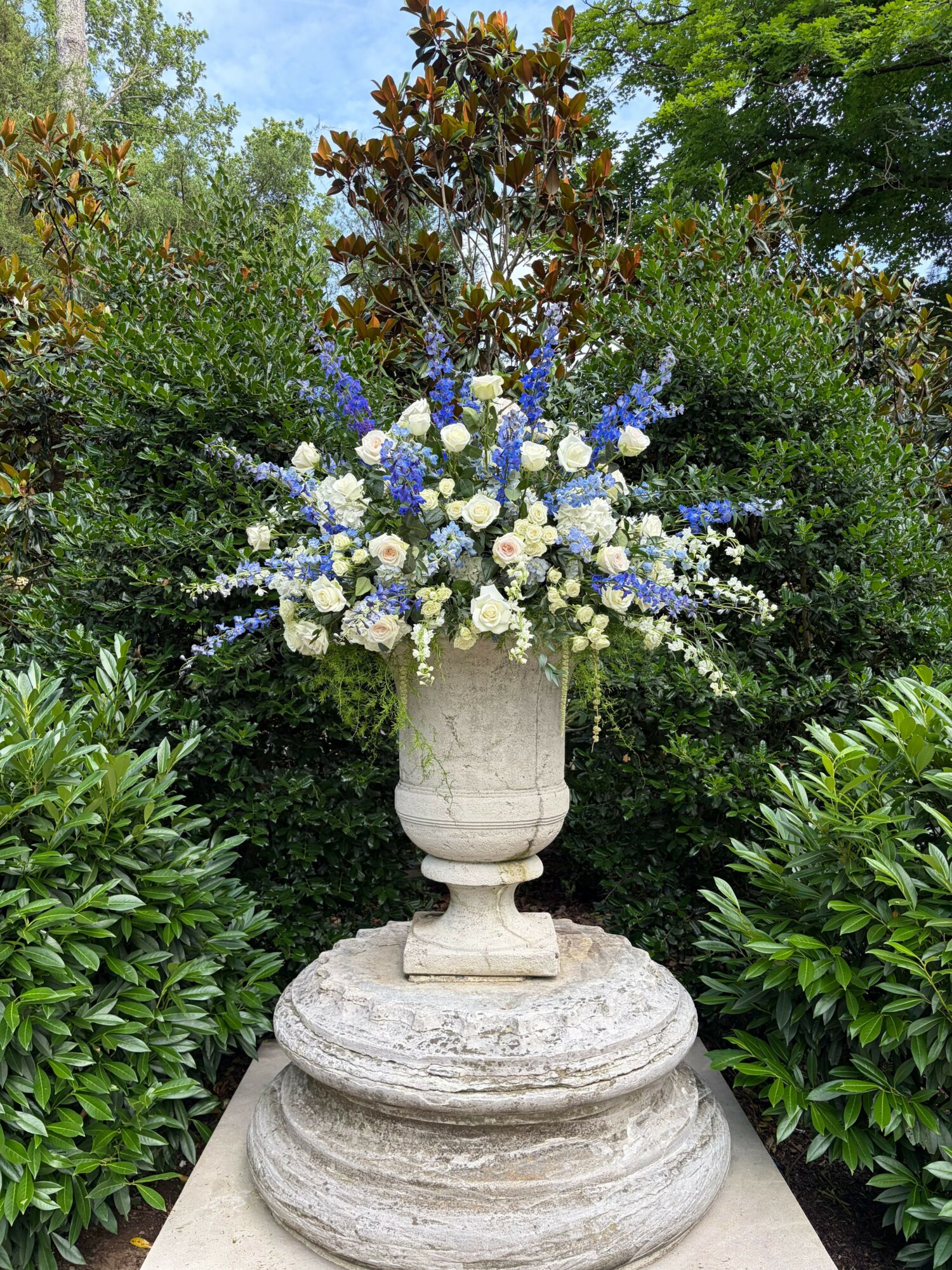Flower arrangement with white and blue flowers in a stone urn on a pedestal, surrounded by green plants and trees.