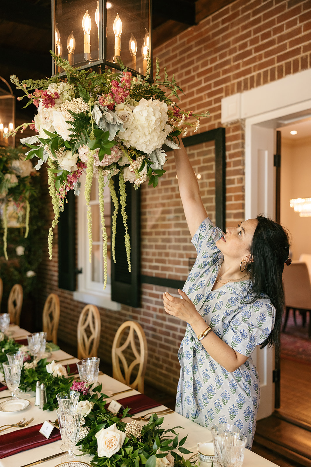 Woman in patterned dress reaching for a large floral arrangement hanging from a chandelier, brick wall background.