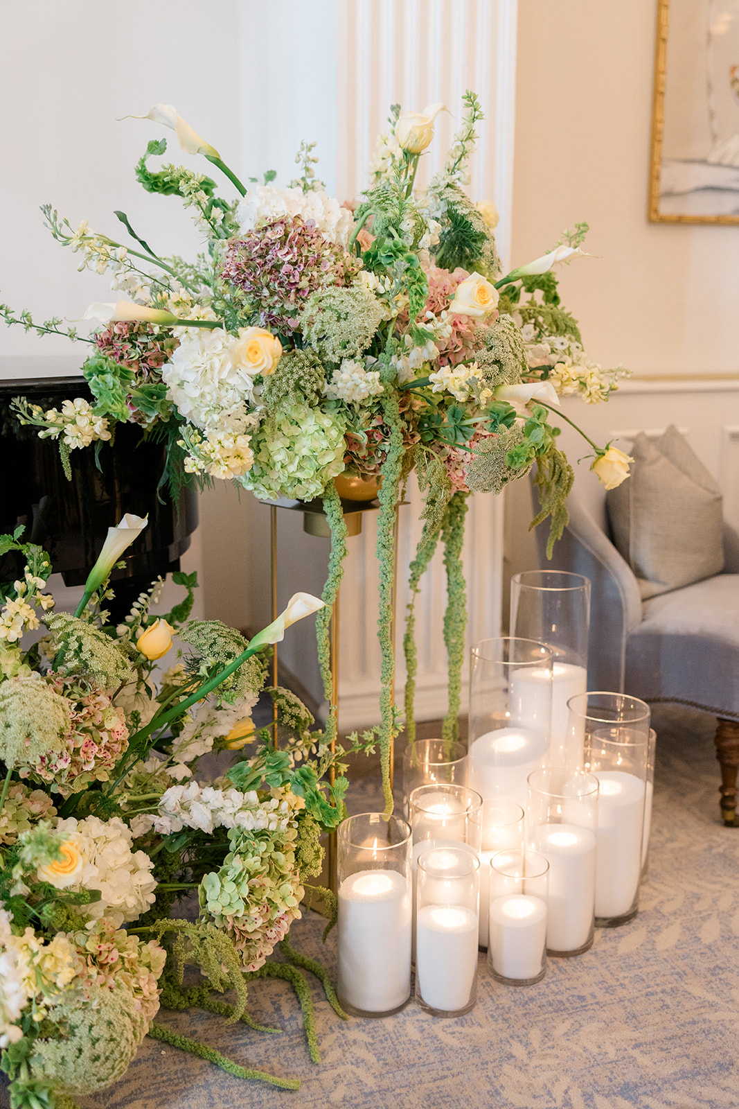 Elegant floral arrangement with white, green, and purple flowers on tall gold stand, surrounded by candles in glass holders, in a decorated room.