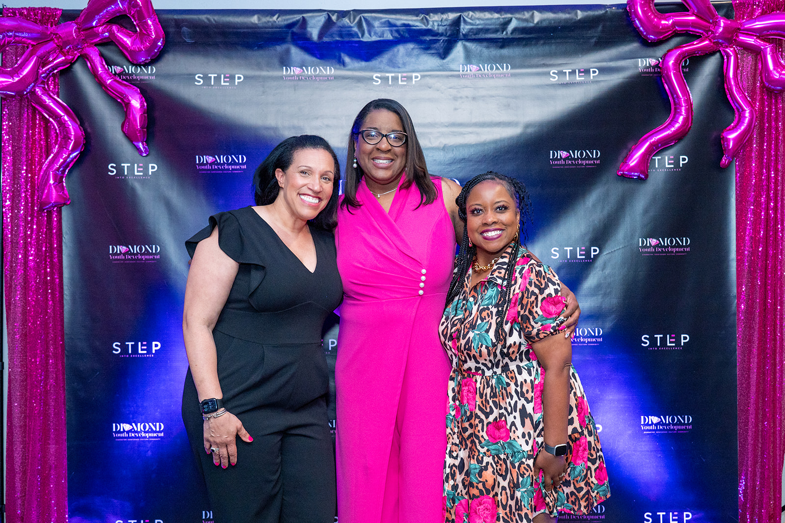 Three women standing together in front of a blue backdrop with pink balloon decorations, smiling for the photo.