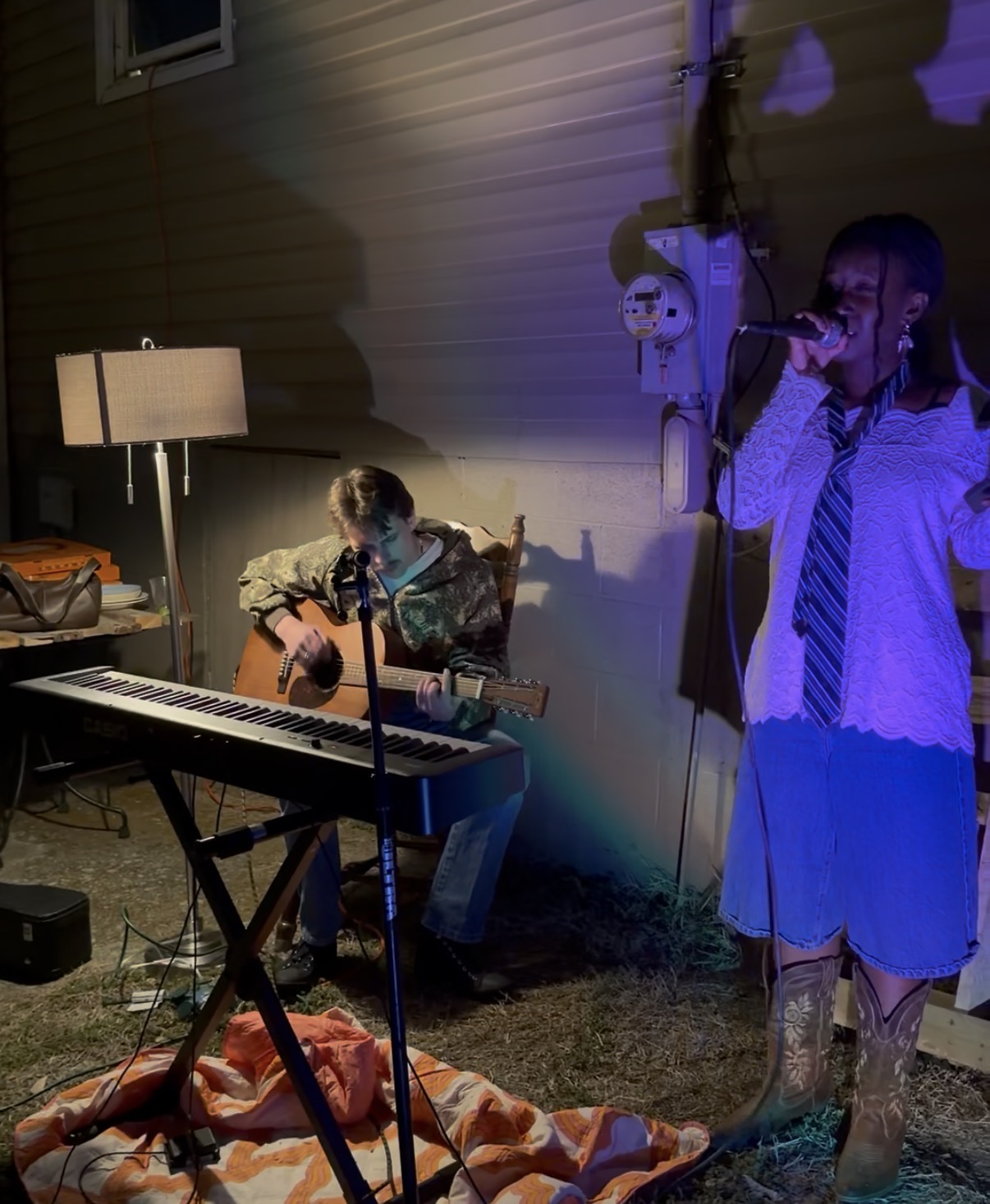 Two people performing music outdoors at night, one playing guitar and the other singing into a microphone, with a keyboard nearby.