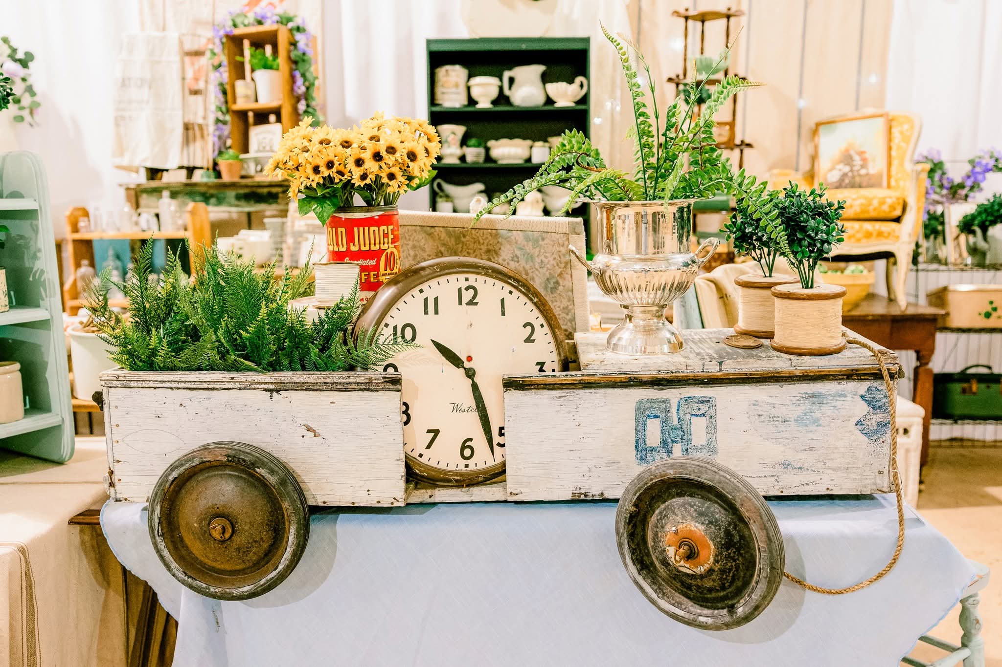 Vintage wooden cart with wheels, filled with plants and a clock, in a room with shelves and flowers.