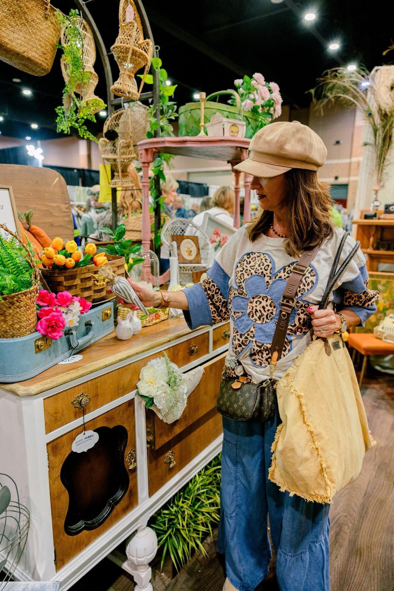 Woman shopping at a store with colorful decorations and flowers, wearing a hat and carrying a yellow bag.