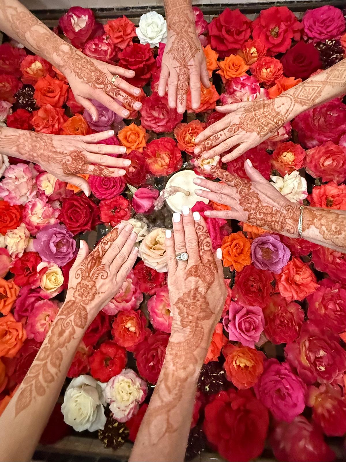 Multiple hands with intricate henna designs reach toward a white object surrounded by colorful roses.