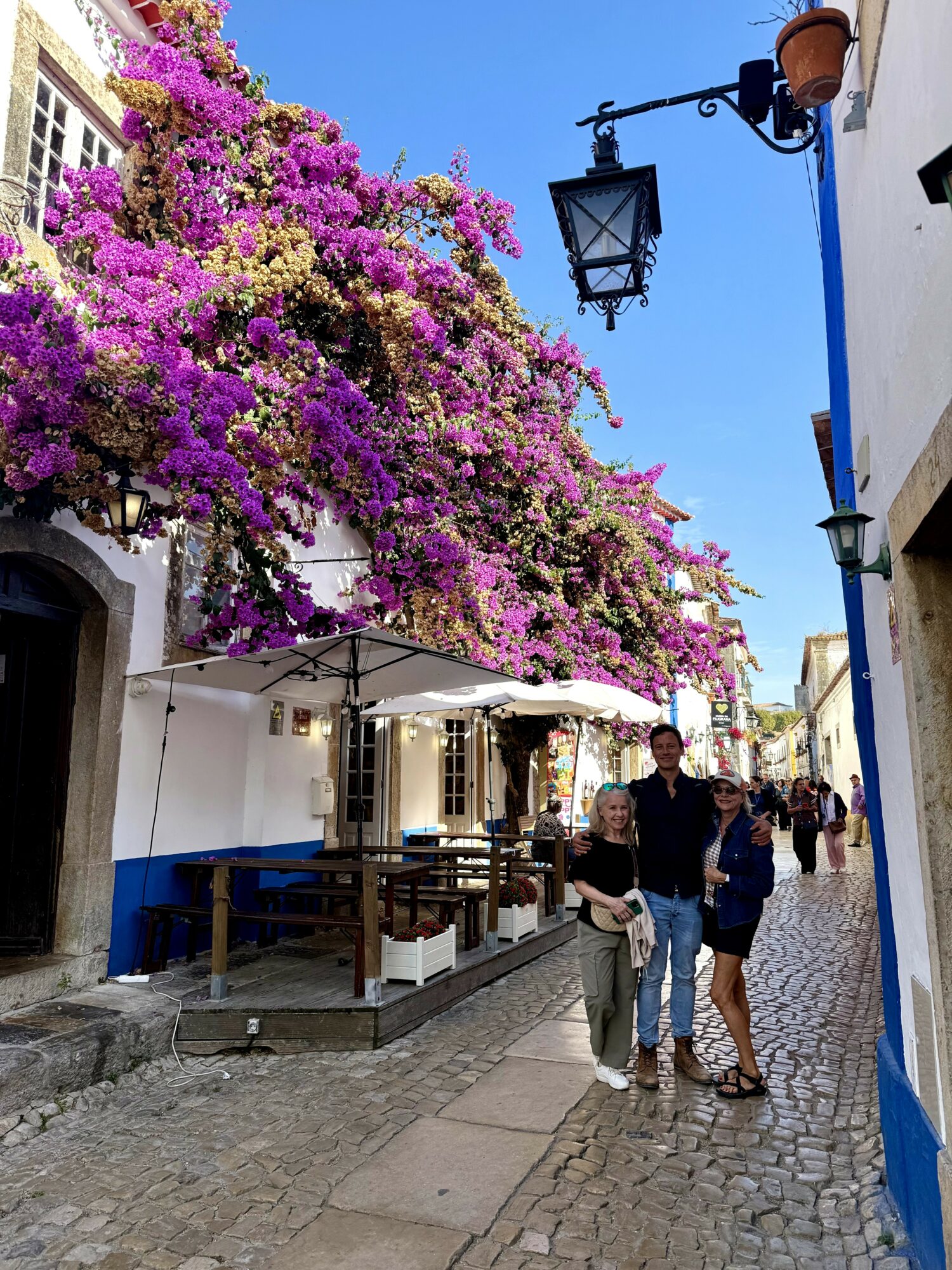 Street scene with white buildings, purple flowering tree, outdoor seating, and three people walking and talking.