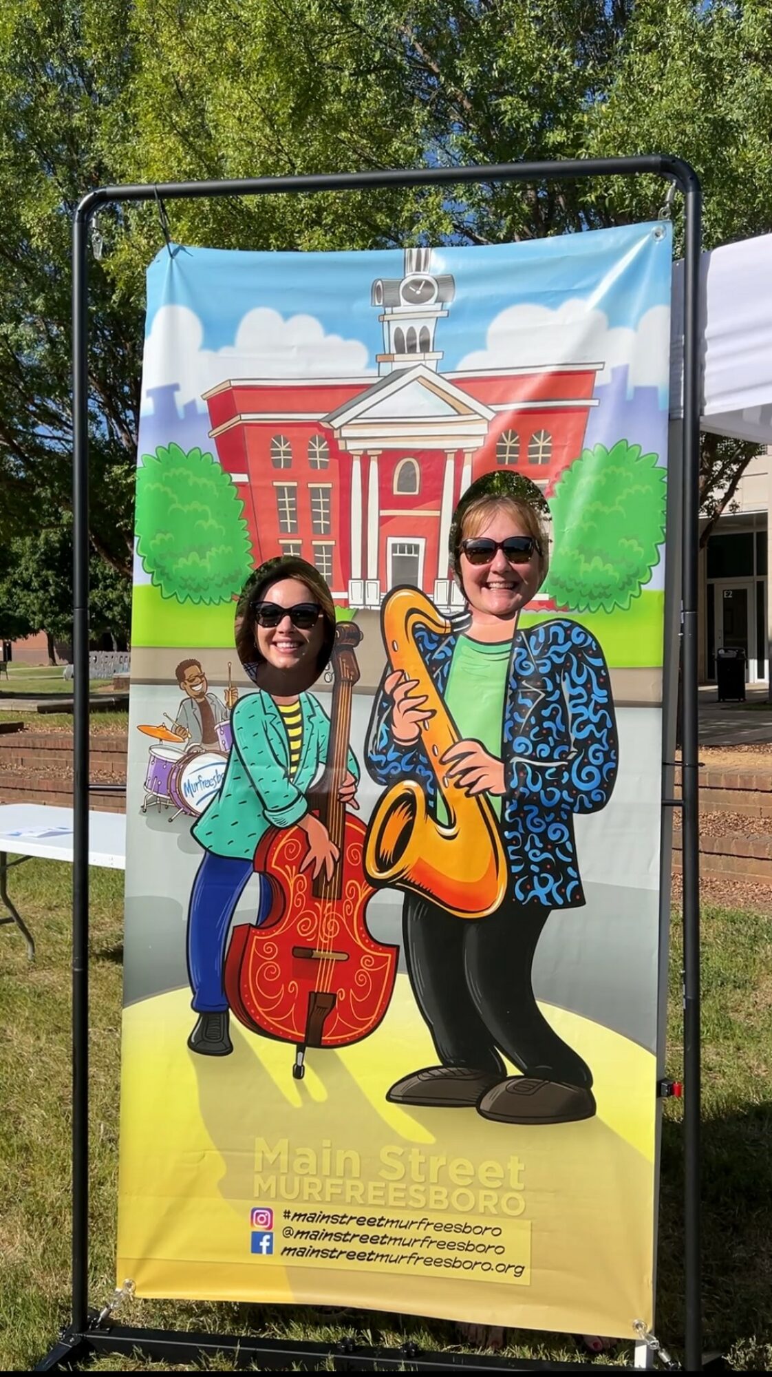 Two women with musical instruments standing in front of a colorful illustrated backdrop of a building and trees.