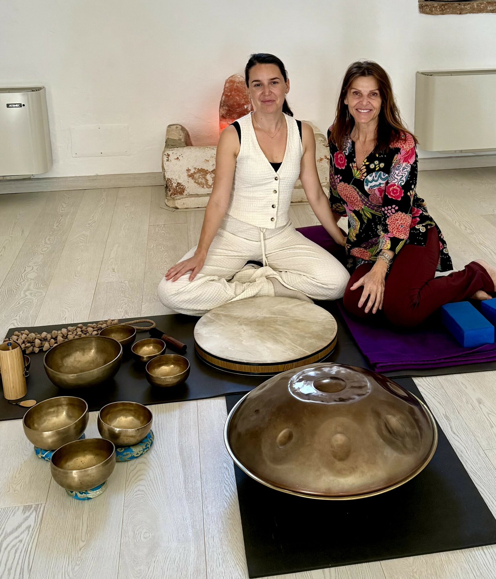 Two women sit on mats with singing bowls and a large handpan instrument in front of them, indoors. One woman wears white, the other colorful clothing.