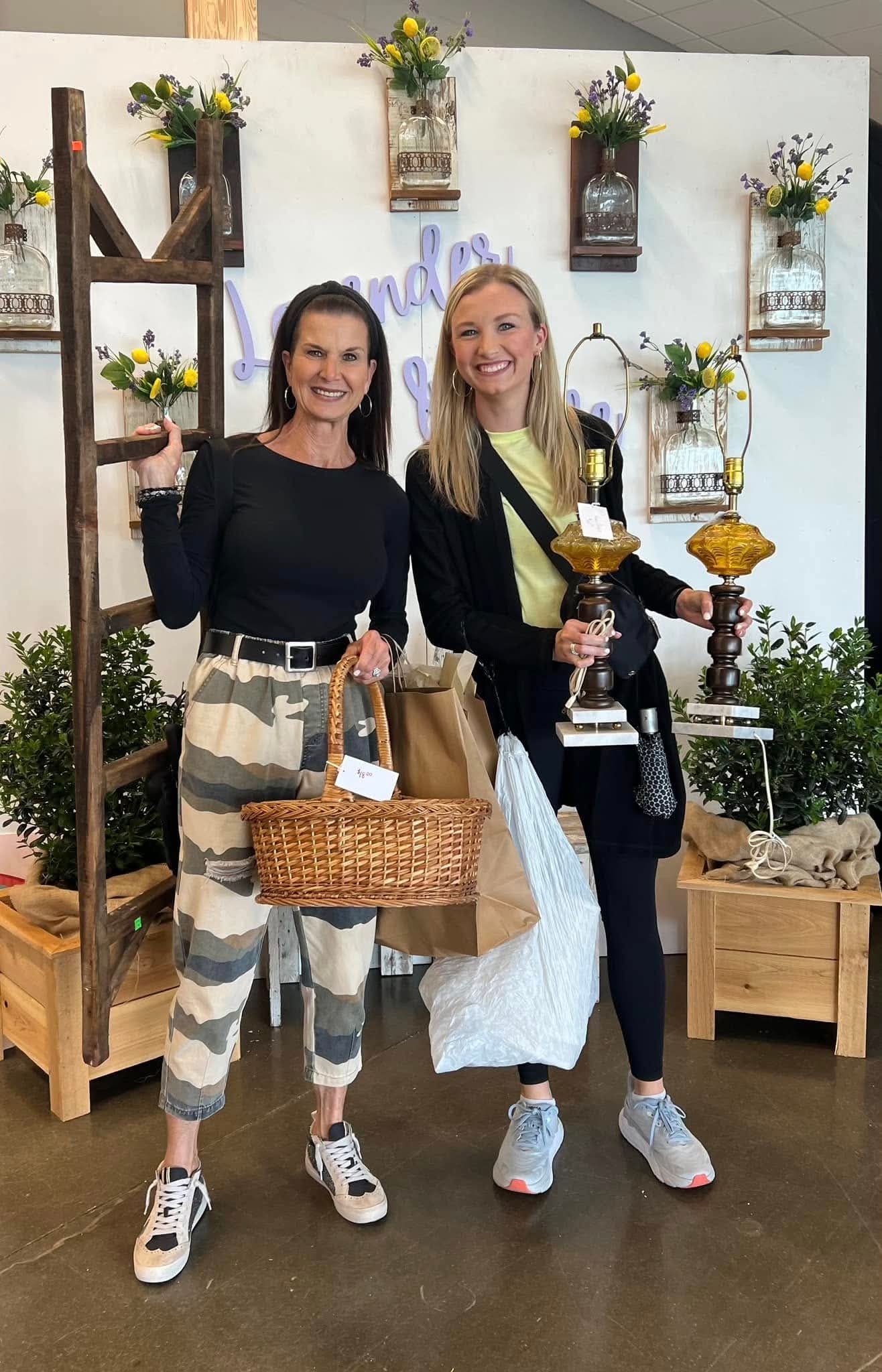 Two women standing indoors, smiling, holding trophies and a basket, with a decorated white wall and plants behind them.