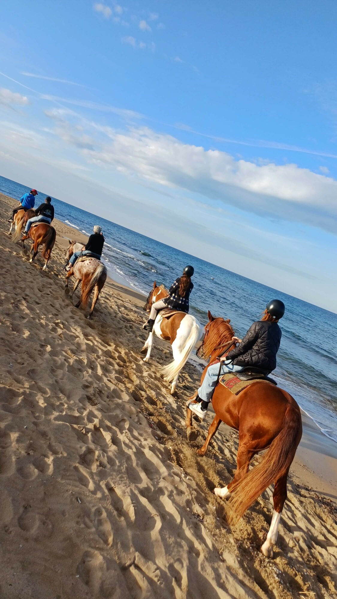 Group of people riding horses along a sandy beach with ocean and sky in background.