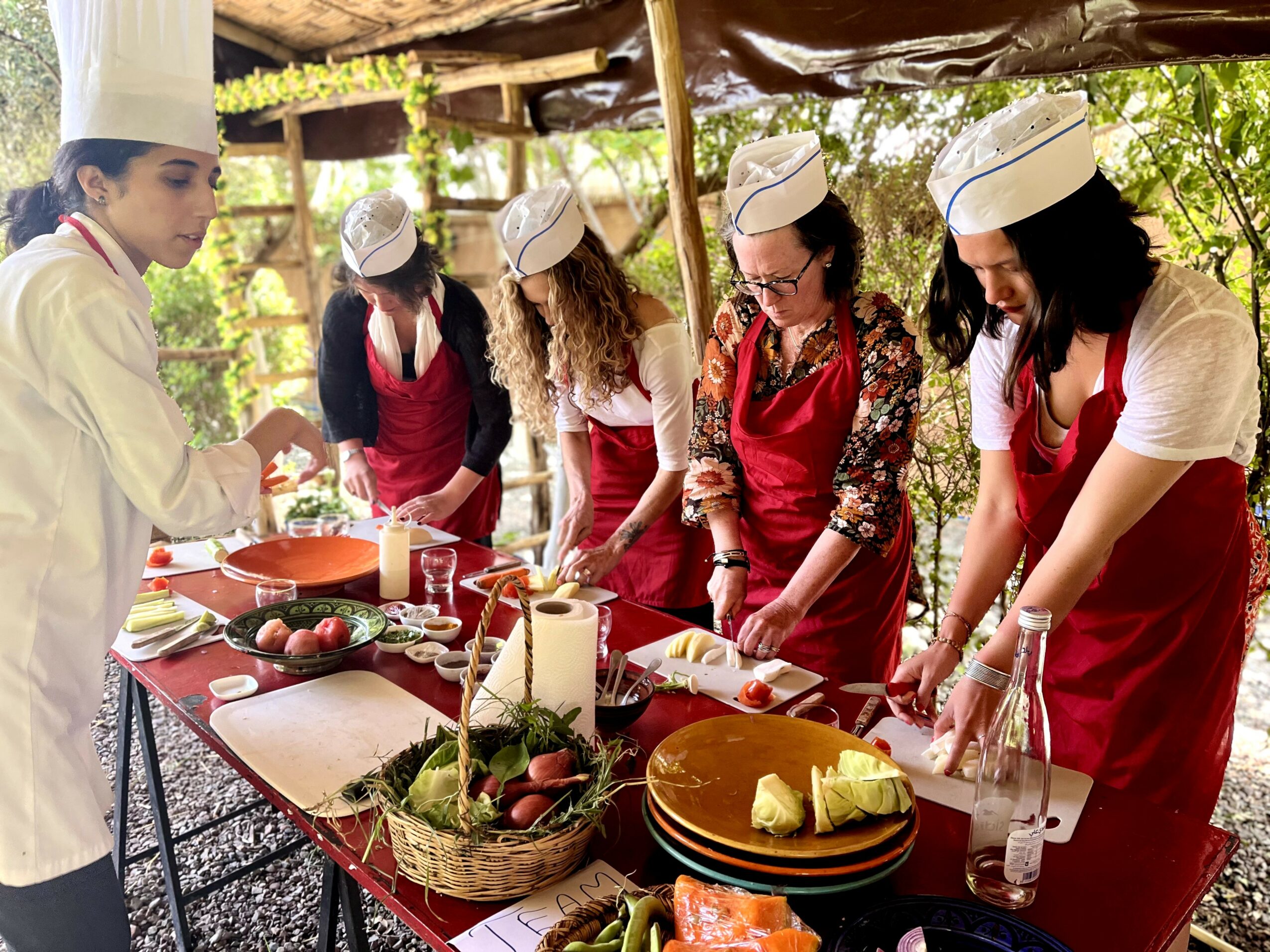 Group of women in chef hats and aprons preparing food outdoors under a wooden shelter, with ingredients and utensils on a table.