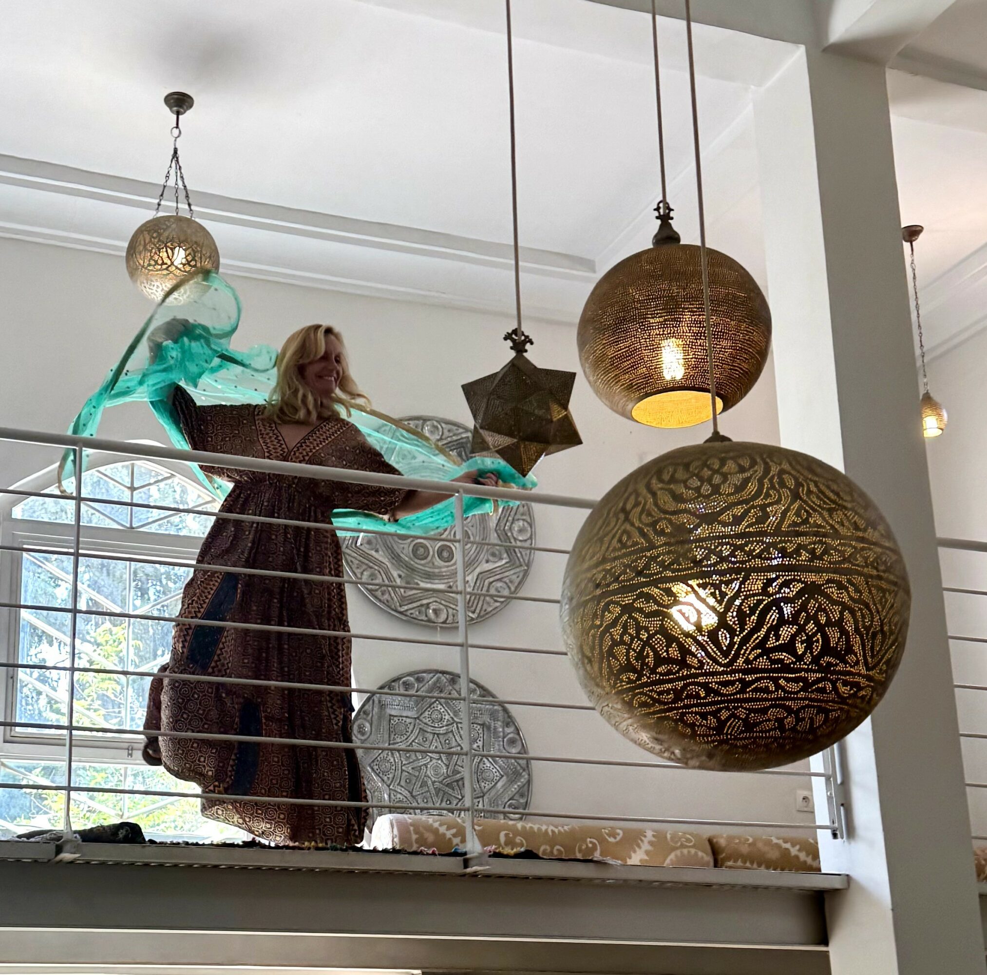 View of hanging decorative lamps and a woman with a flowing scarf near a window, seen from below.