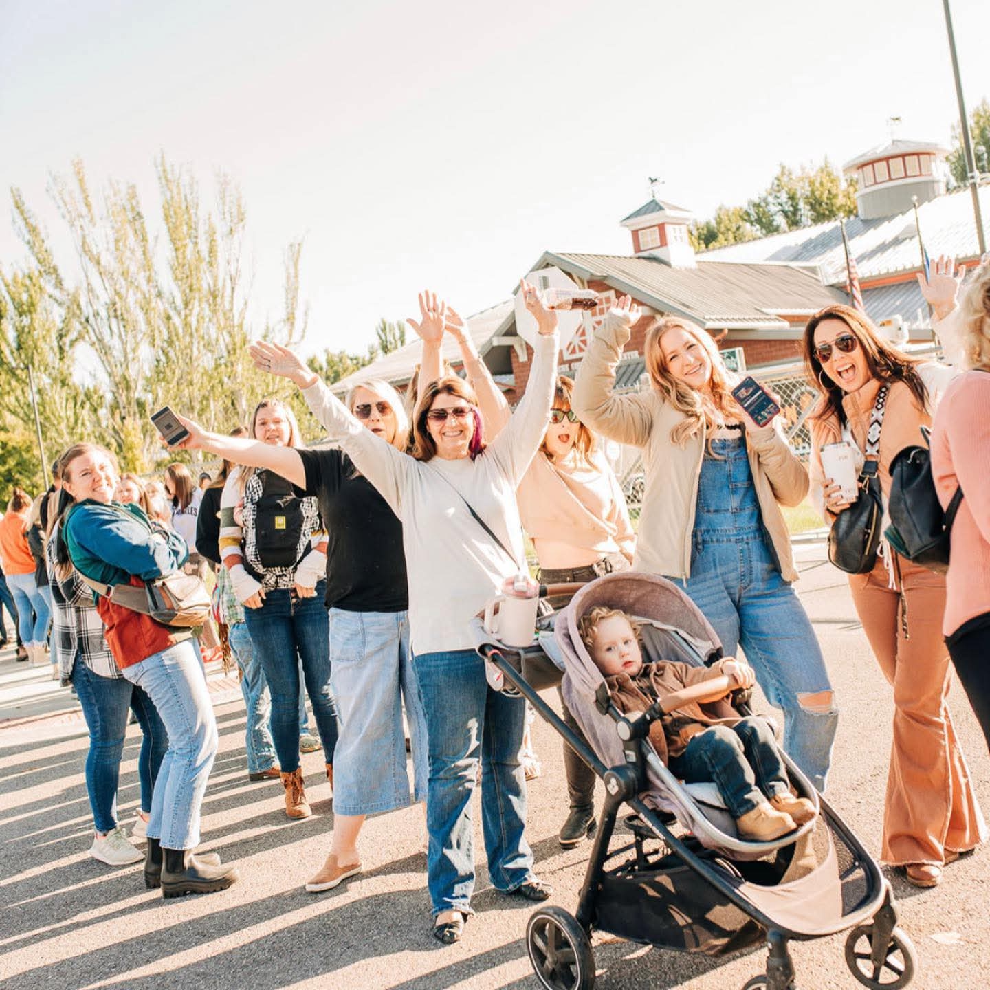 Group of people outdoors, some smiling and waving, with a stroller and a building in the background.