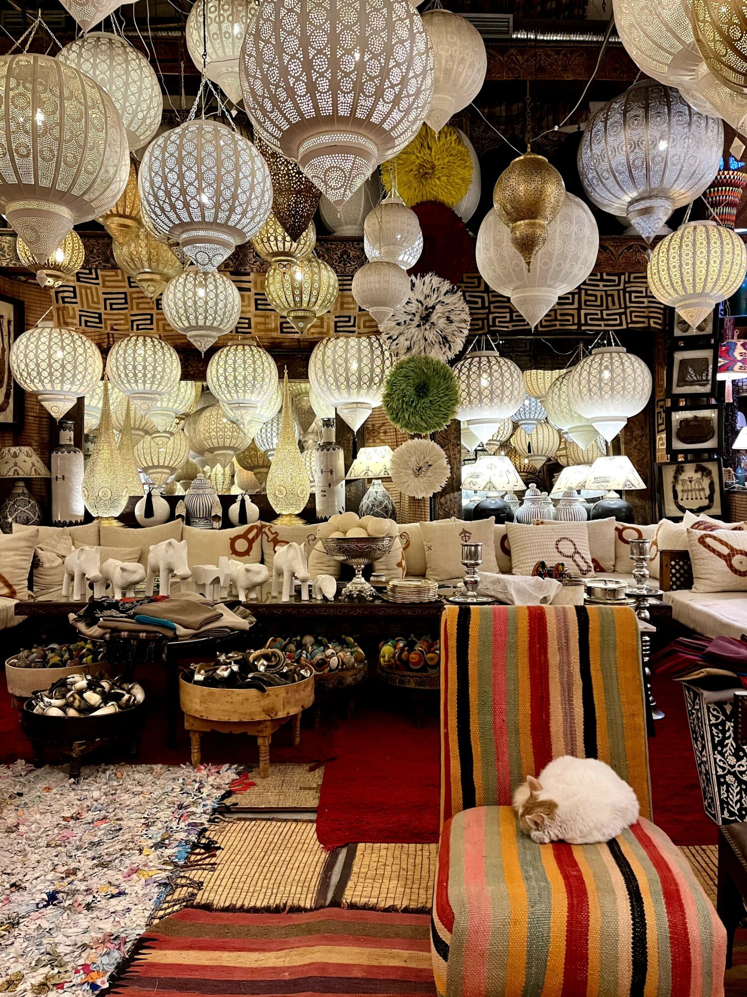 Room decorated with hanging paper lanterns, a sofa with cushions, a striped chair with a cat, and colorful rugs.