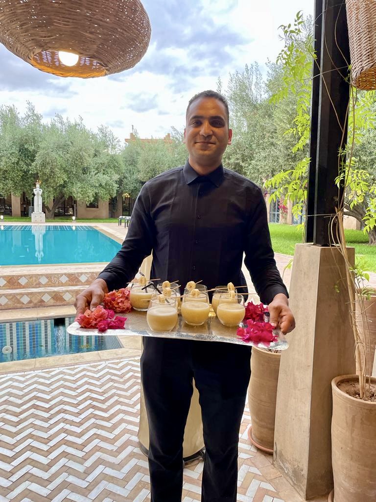Man in black shirt holding a tray of drinks and flowers outdoors near a pool, with trees and sky in background.