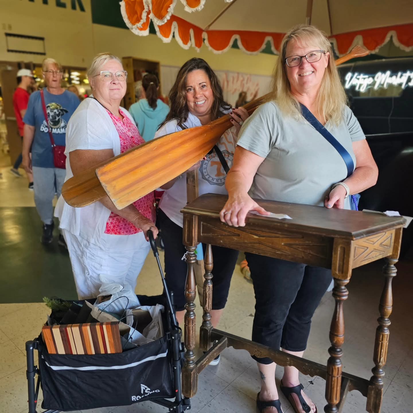 Three women standing indoors, one with a wooden paddle, smiling, with a table and shopping cart nearby.