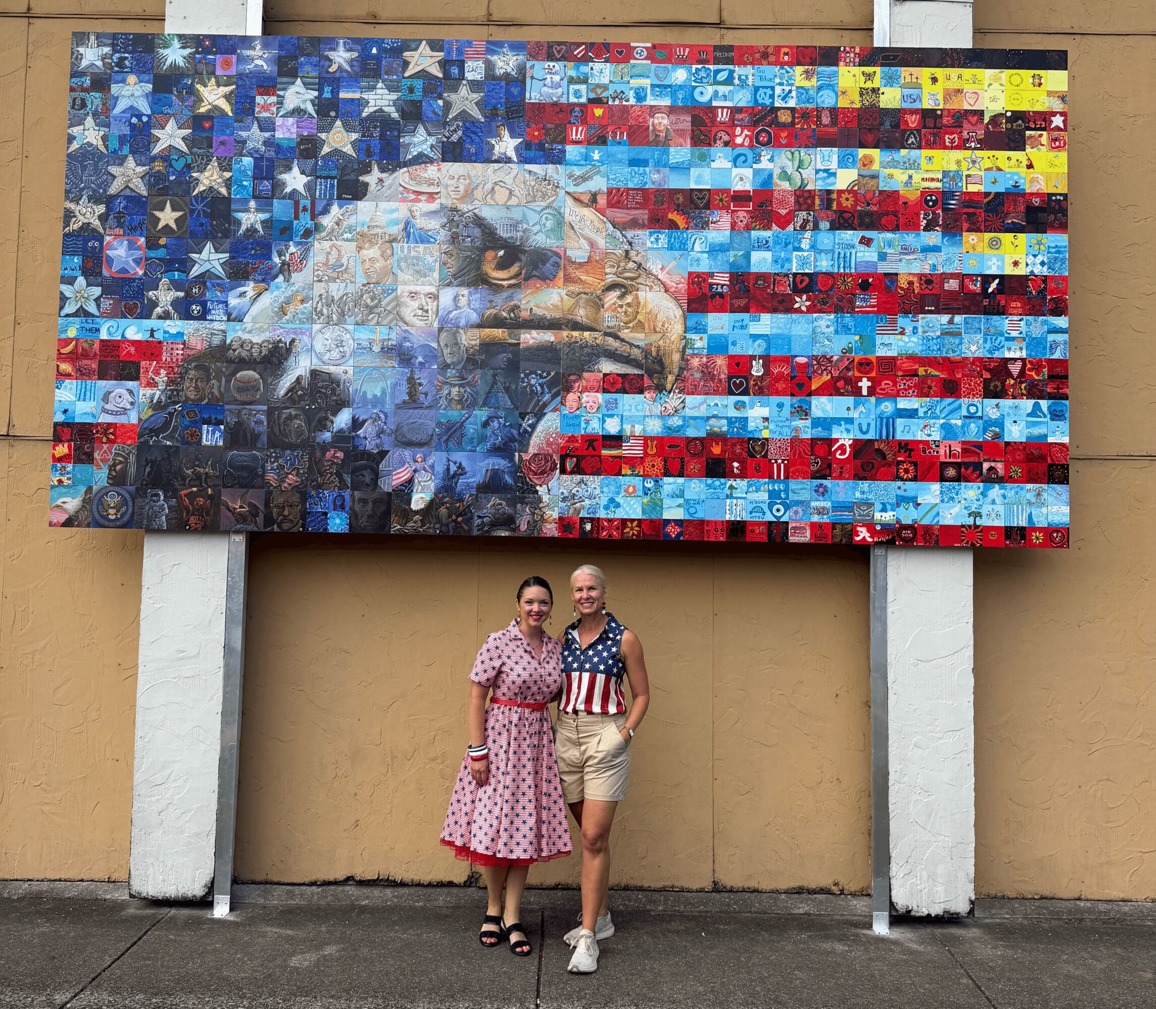 Two women stand in front of a large mosaic mural of an eagle with an American flag background, smiling.