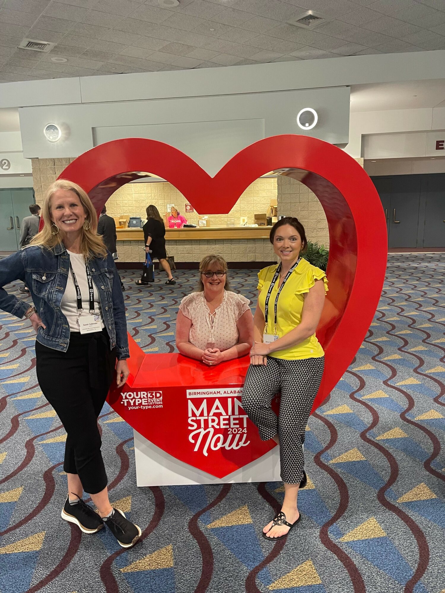 Three women sitting on a red heart-shaped bench with a sign, in an indoor setting, smiling at the camera.