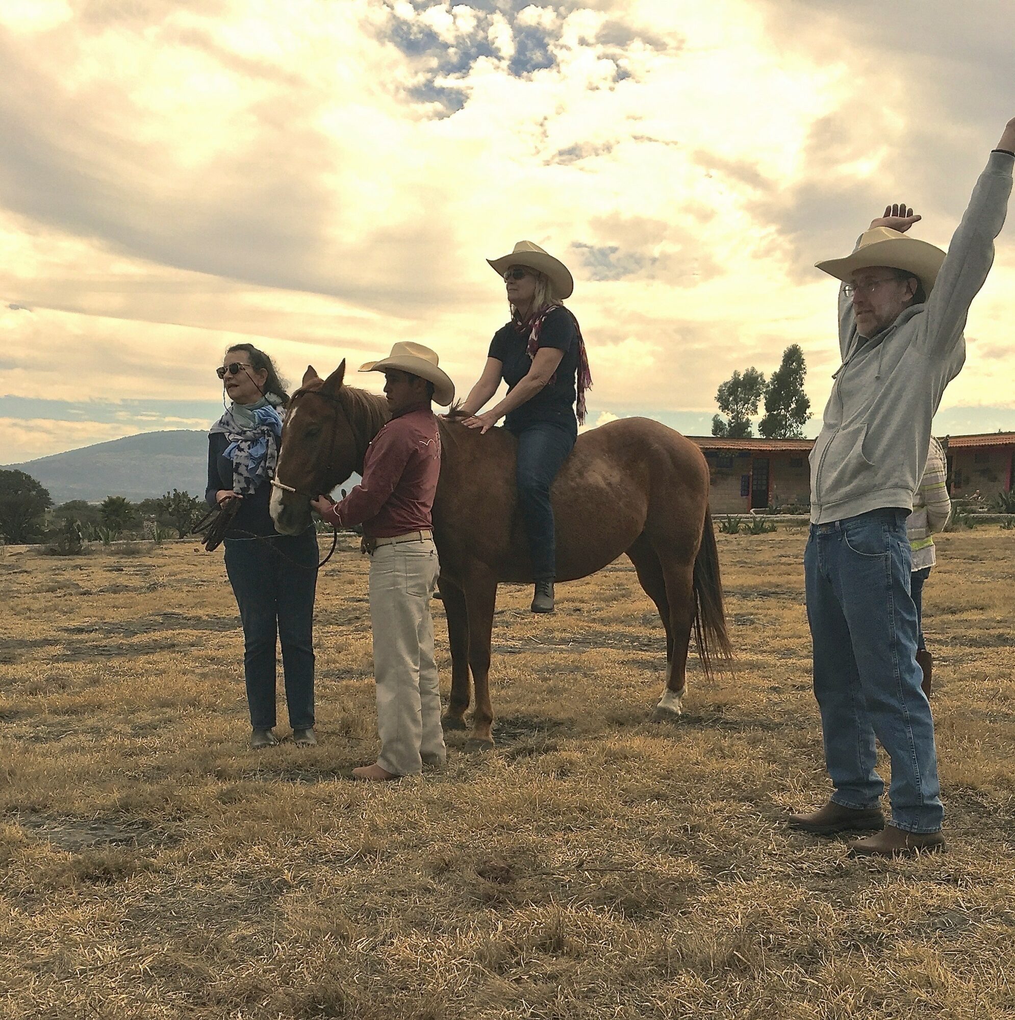 Four people, two children and two adults, stand outdoors with a horse, one child is riding the horse, and the others are standing nearby, under a cloudy sky.