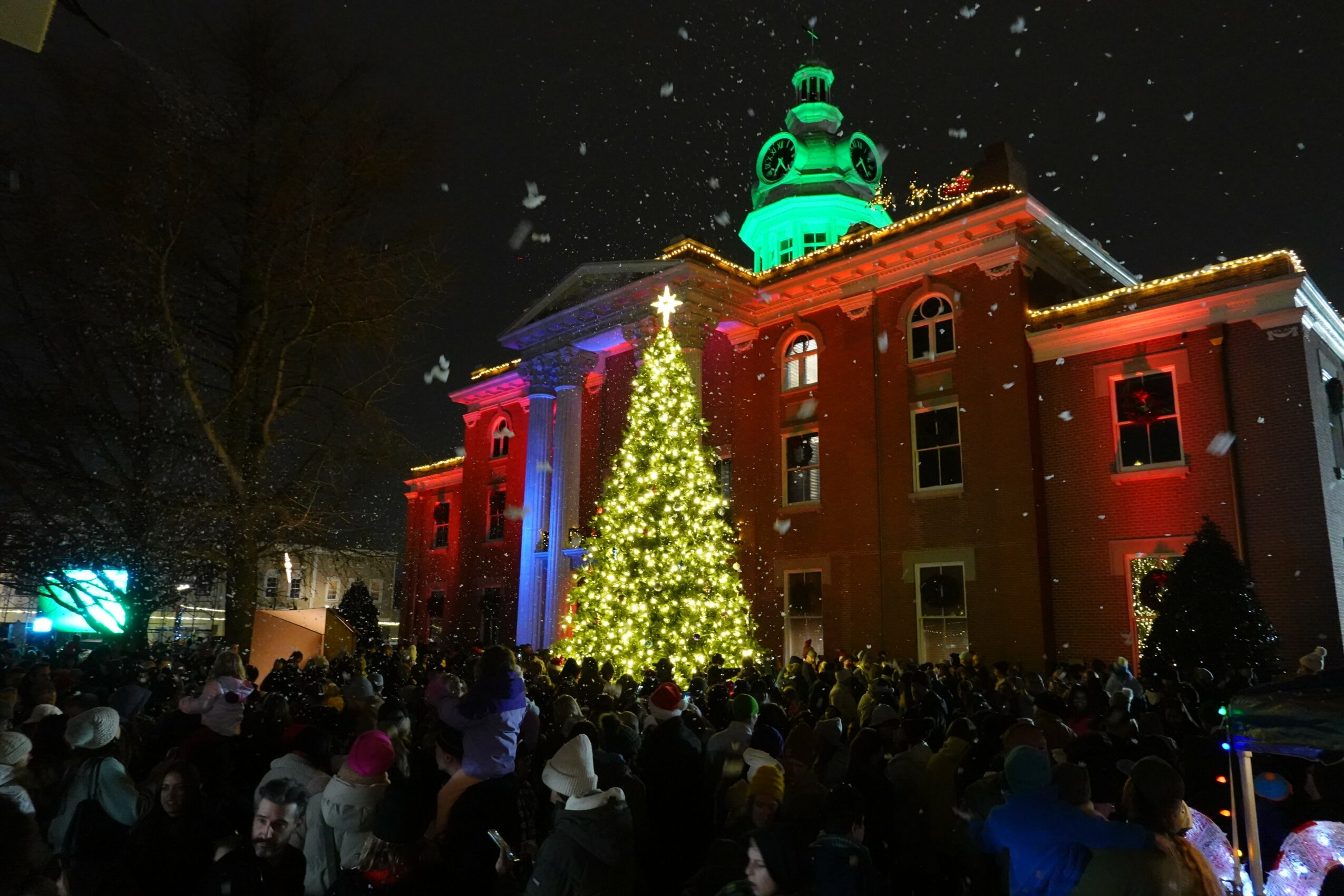 A large crowd gathers outside a decorated building with a Christmas tree, illuminated with colorful lights at night.