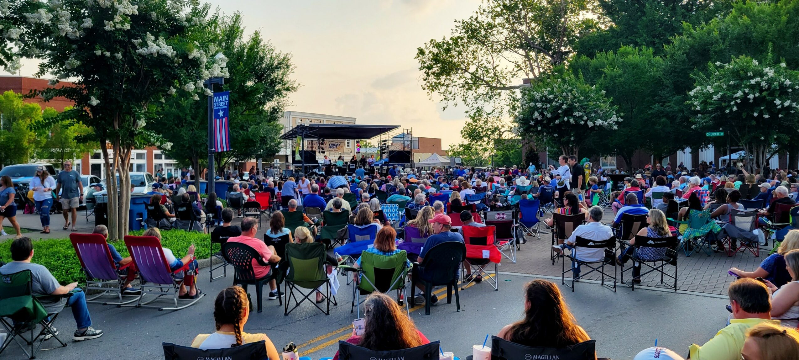 Crowd gathered outdoors with chairs and tables, trees and buildings in the background, people seated and socializing.