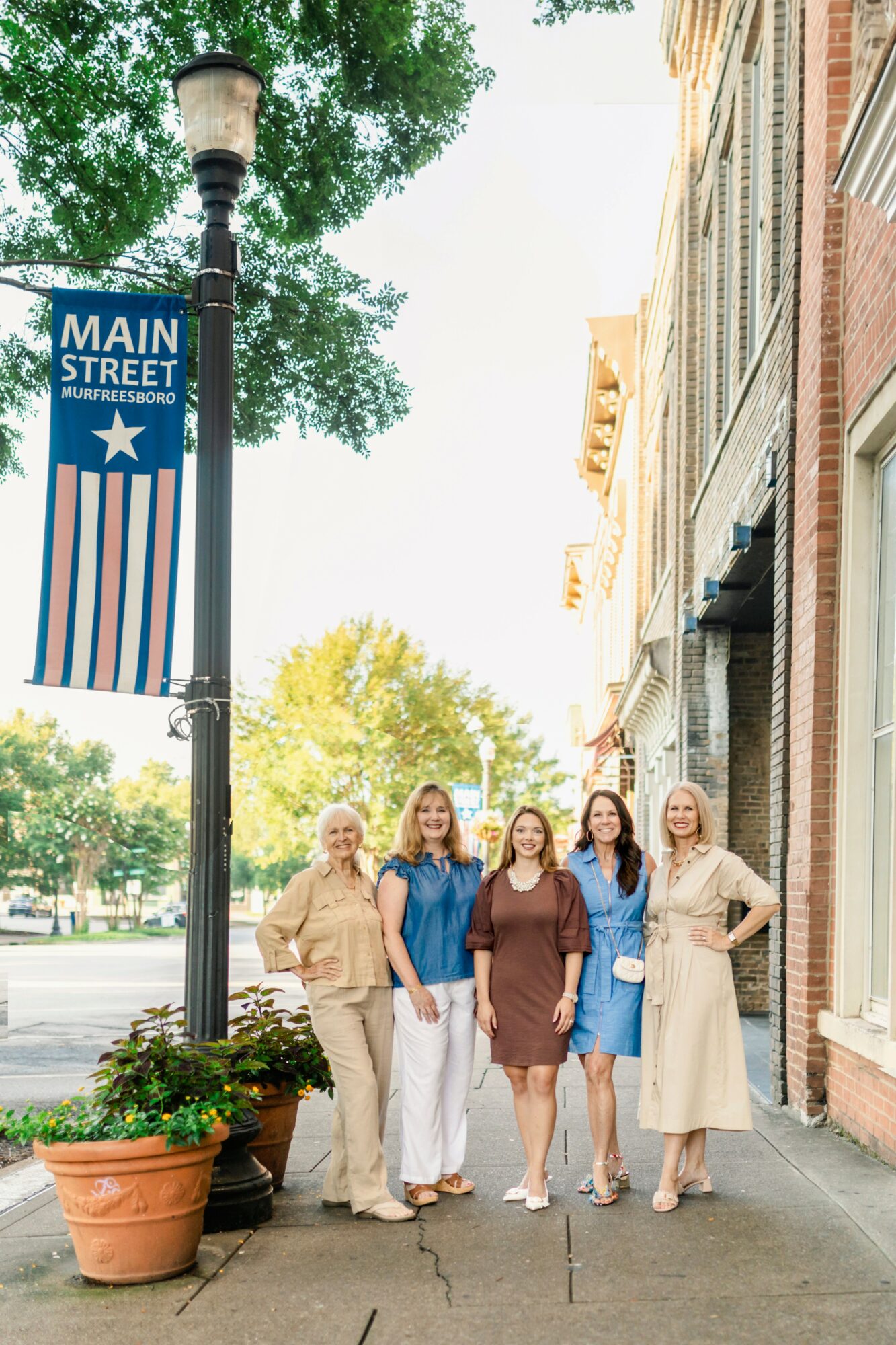 Five women stand on a sidewalk near a streetlamp and a building, smiling at the camera, with trees and buildings in the background.