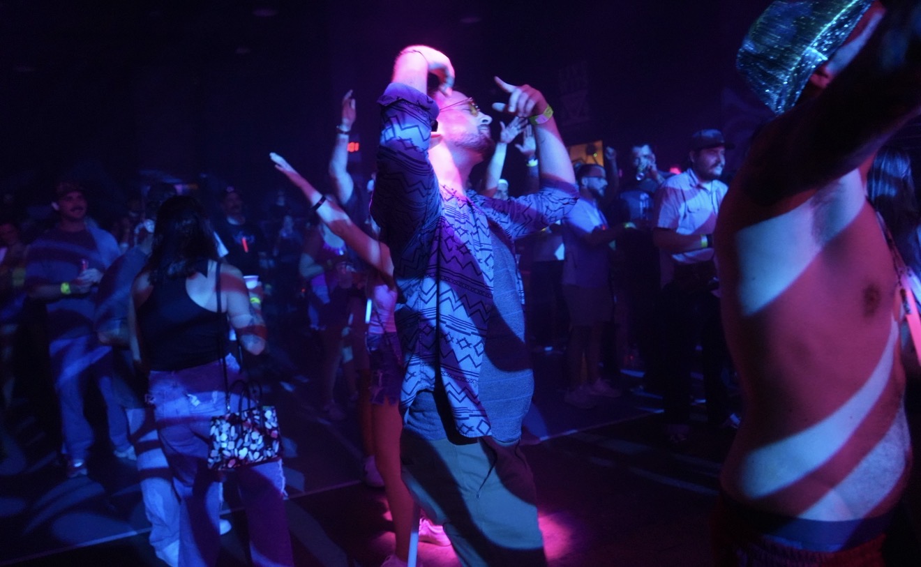 People dancing in a dark, colorful club with blue and purple lighting, some with arms raised, enjoying music.