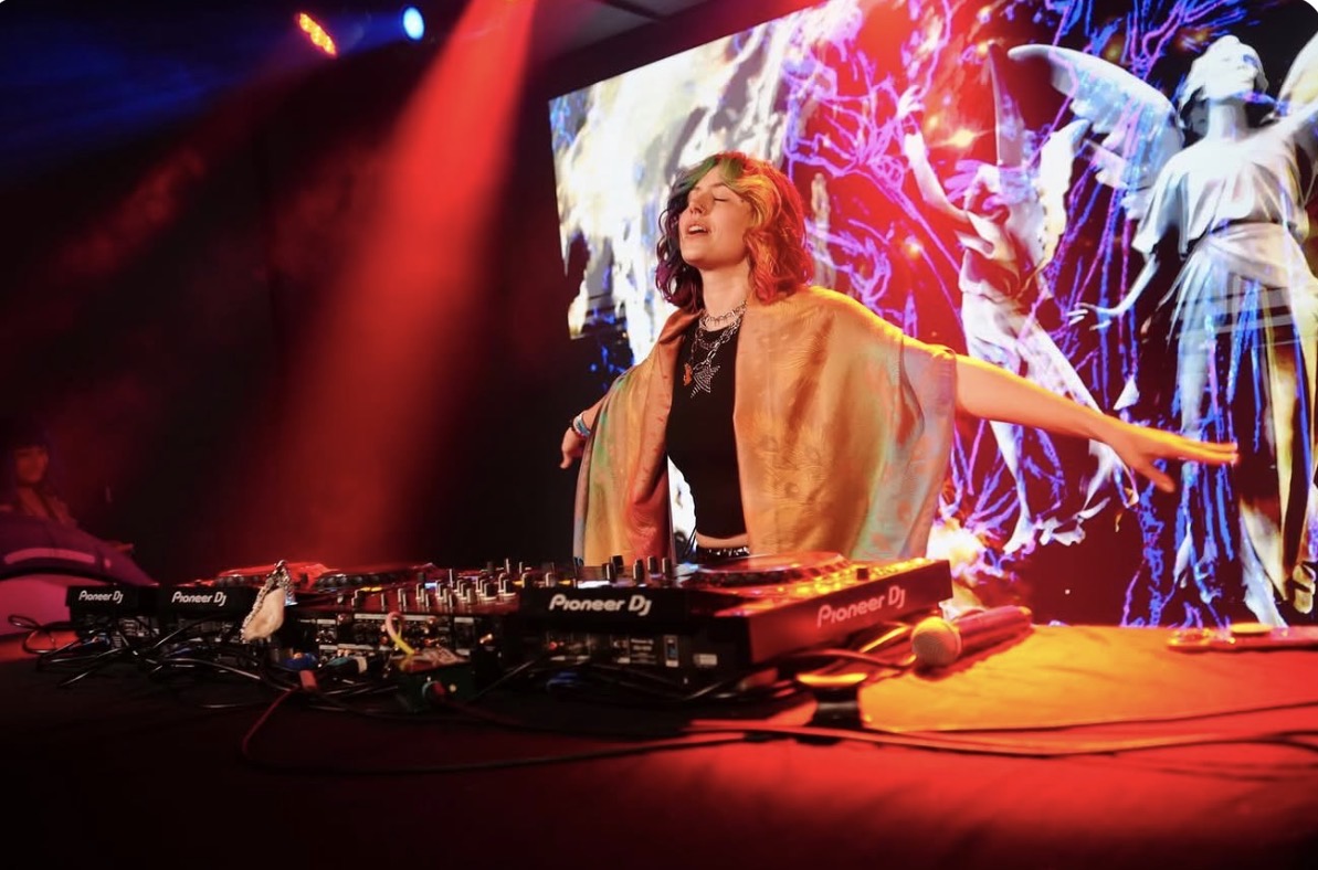 DJ woman with arms outstretched performing on stage with colorful background and dancers, red lighting, DJ equipment in front.