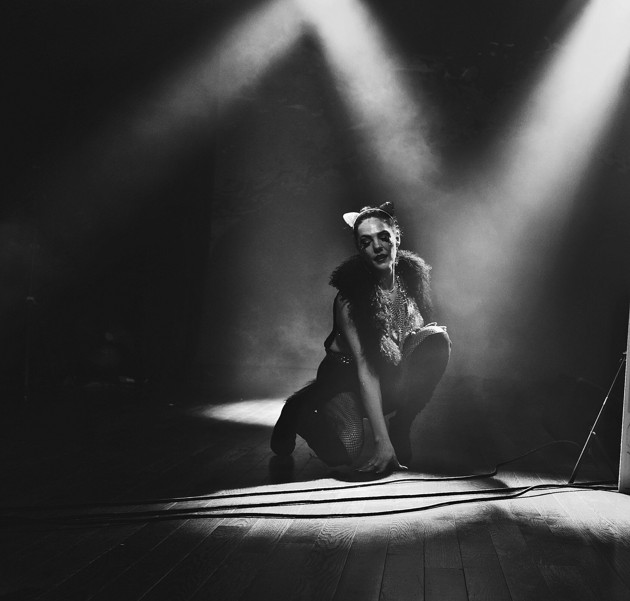 Person crouching on stage with cat ears, illuminated by spotlights, in a dark setting.