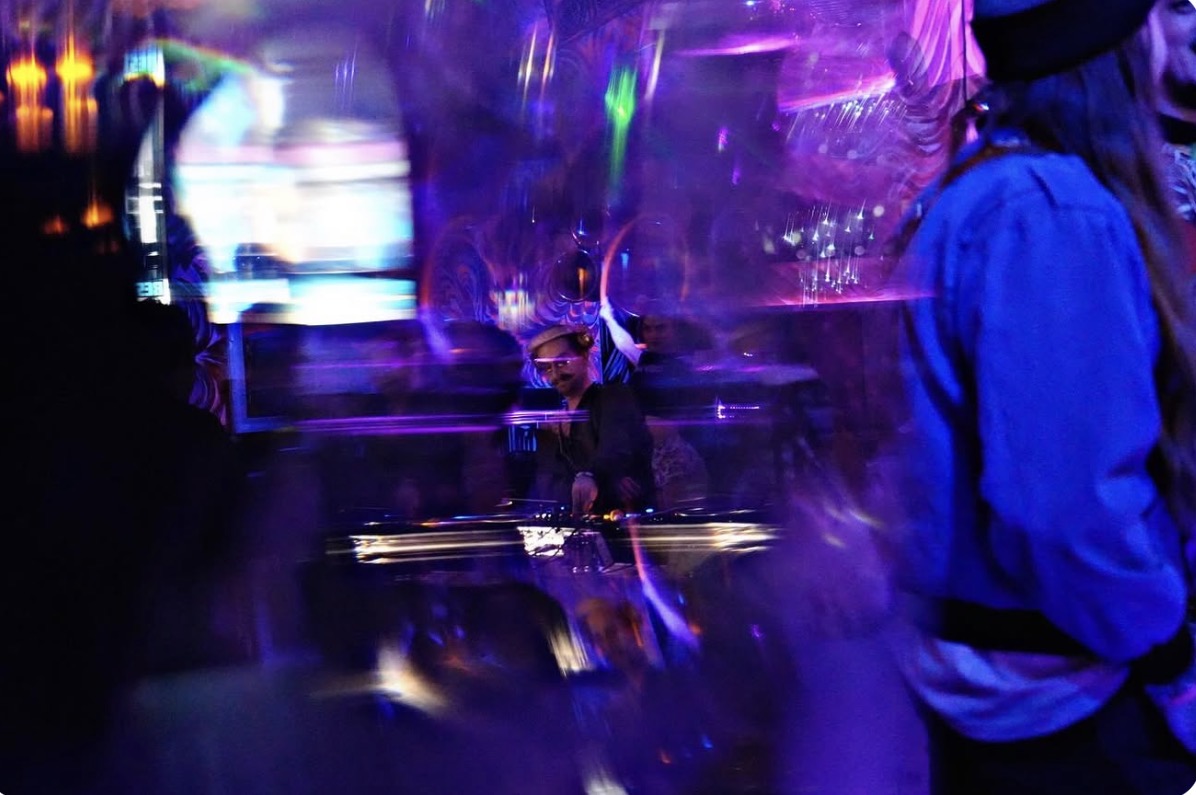 Person in a blue jacket and hat in a colorful, blurry environment with purple and blue lights.
