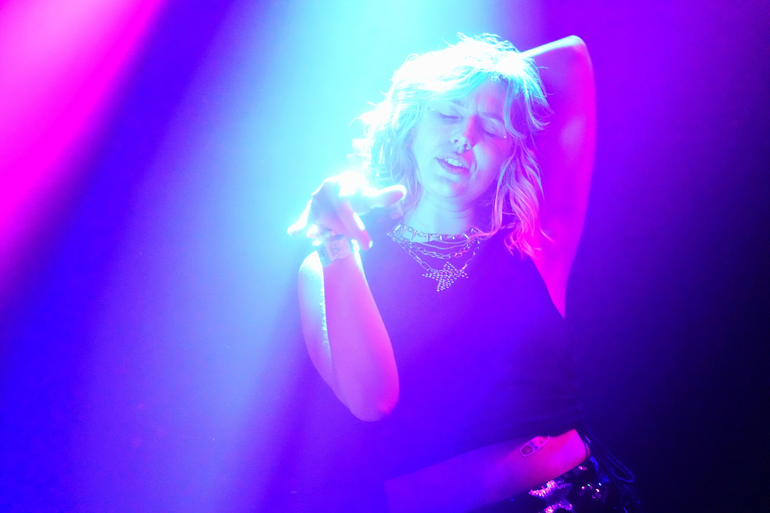 Woman with wavy hair in colorful lighting, eyes closed, posing with one arm raised, wearing a necklace.