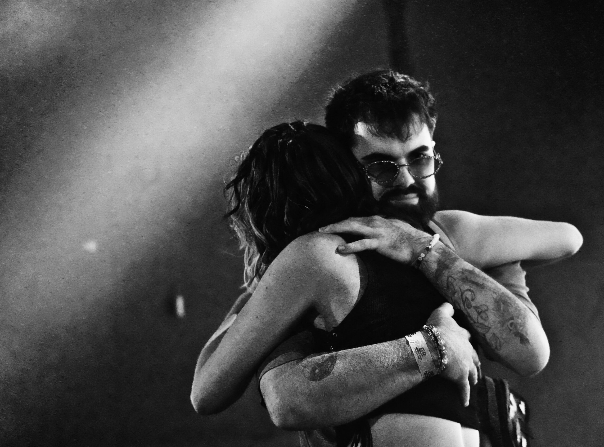 Two people hugging, one with glasses and tattoos, in a black and white photo.