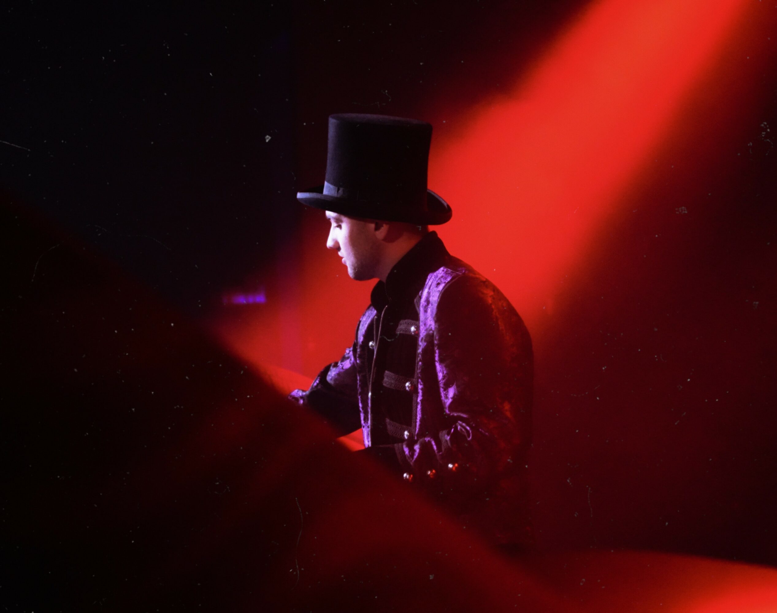 Person wearing a top hat and velvet jacket, illuminated by red stage lighting, performing on stage.