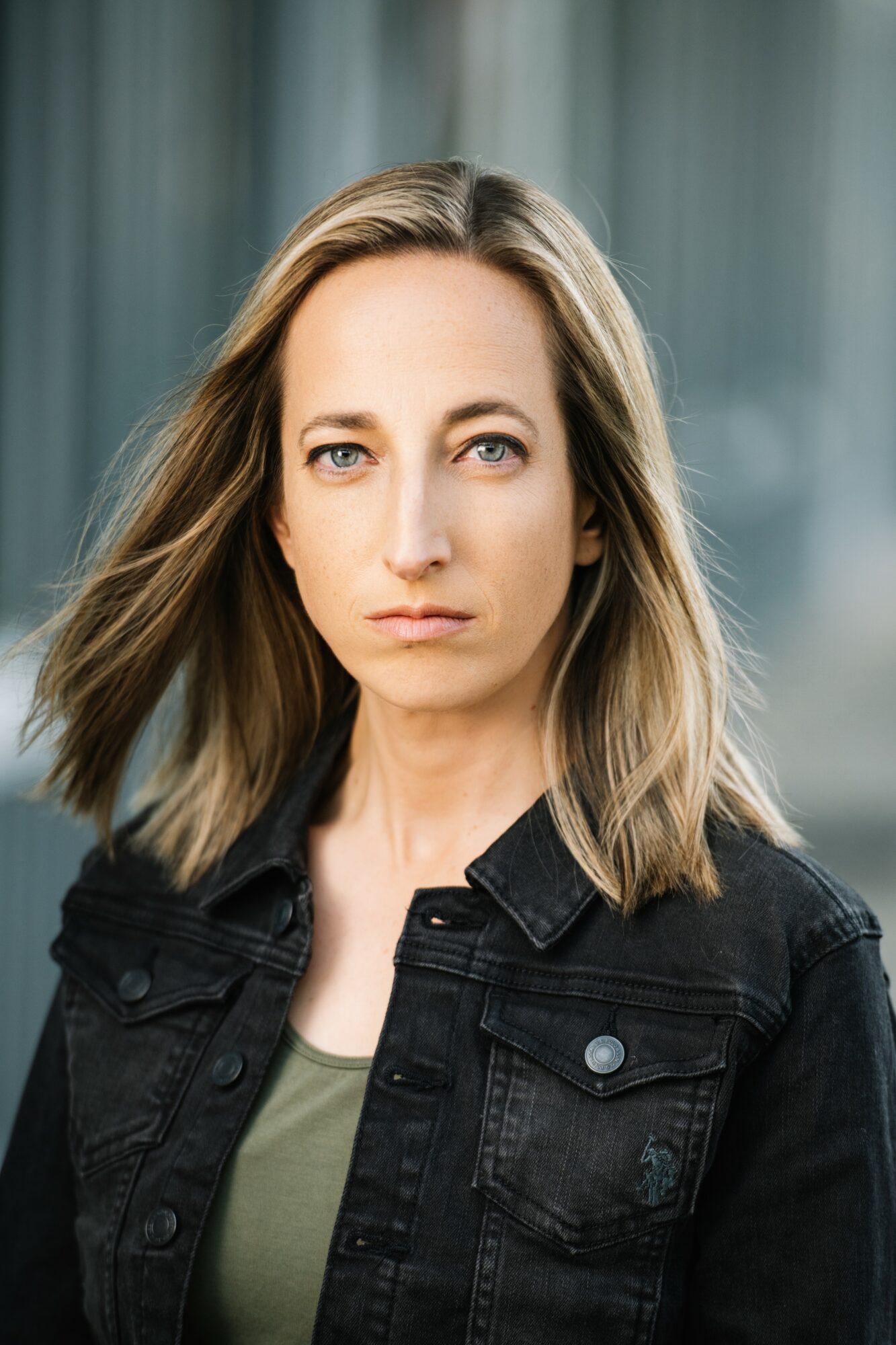 Woman with shoulder-length hair wearing a black jacket and green top, standing outdoors with blurred background.