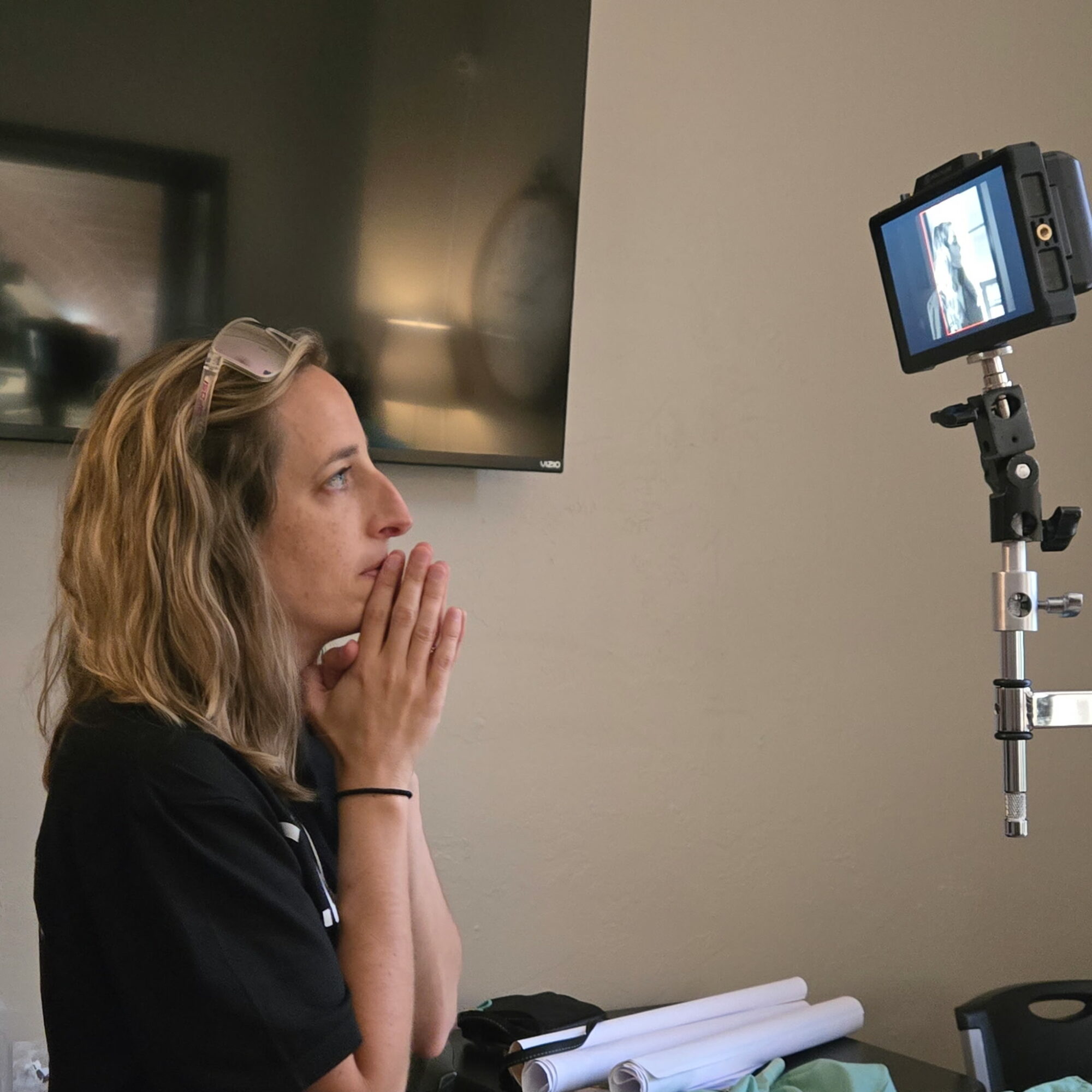 Woman with wavy hair looking at a camera monitor, hands near her face, in a room with a TV and papers.