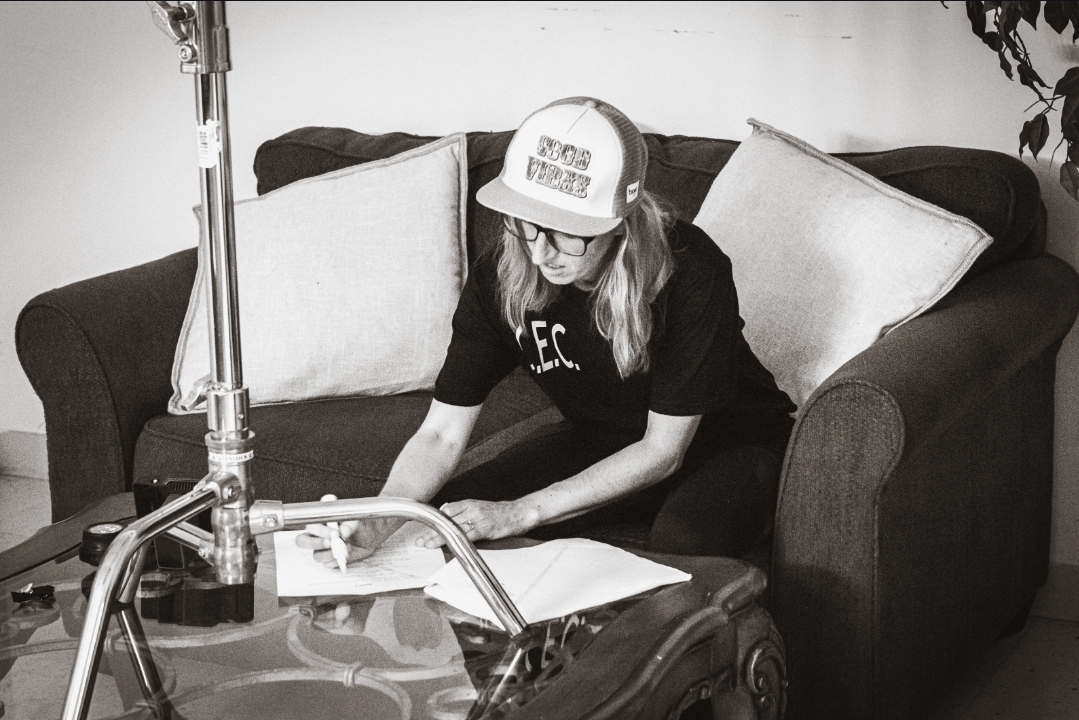 Person sitting on a couch, wearing a cap, writing or drawing on paper, with pillows behind them.