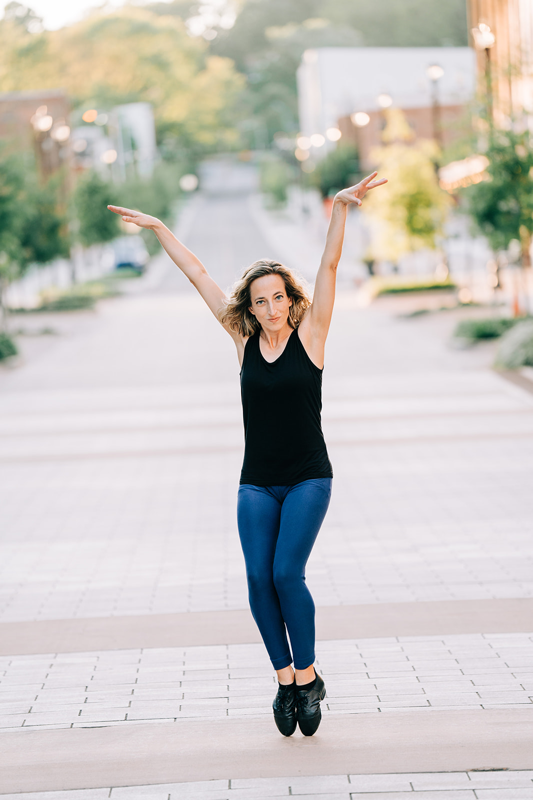 Woman jumping with arms raised on city street, trees and buildings in background, wearing black top and blue jeans.