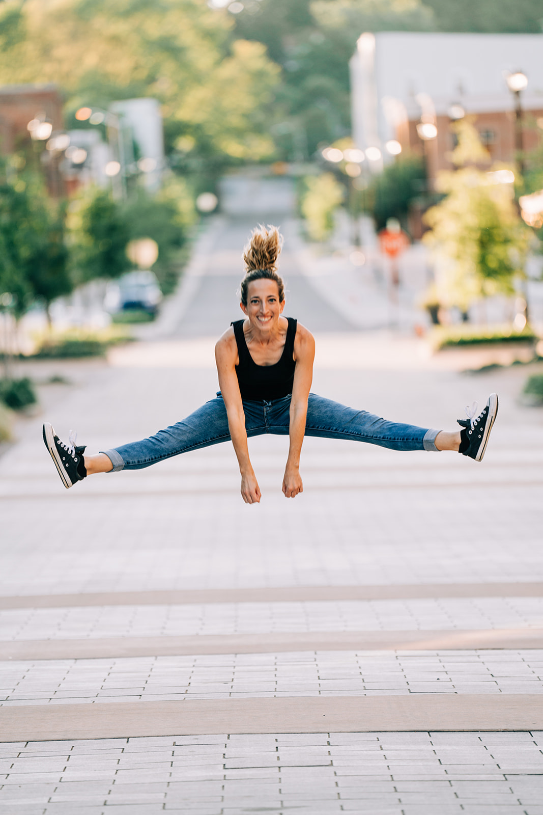 Young woman performing a split jump outdoors on a city street, smiling, with trees and buildings in the background.