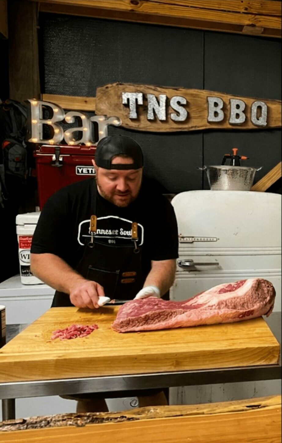 Man preparing meat on a wooden cutting board at a barbecue restaurant, with a sign reading 'TNS BBQ' in the background.