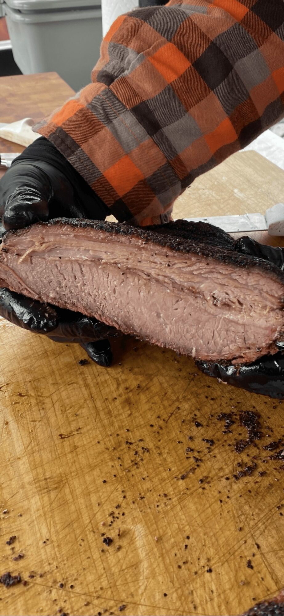 Person in plaid shirt holding a smoked meat cut on a wooden surface, with black gloves visible.