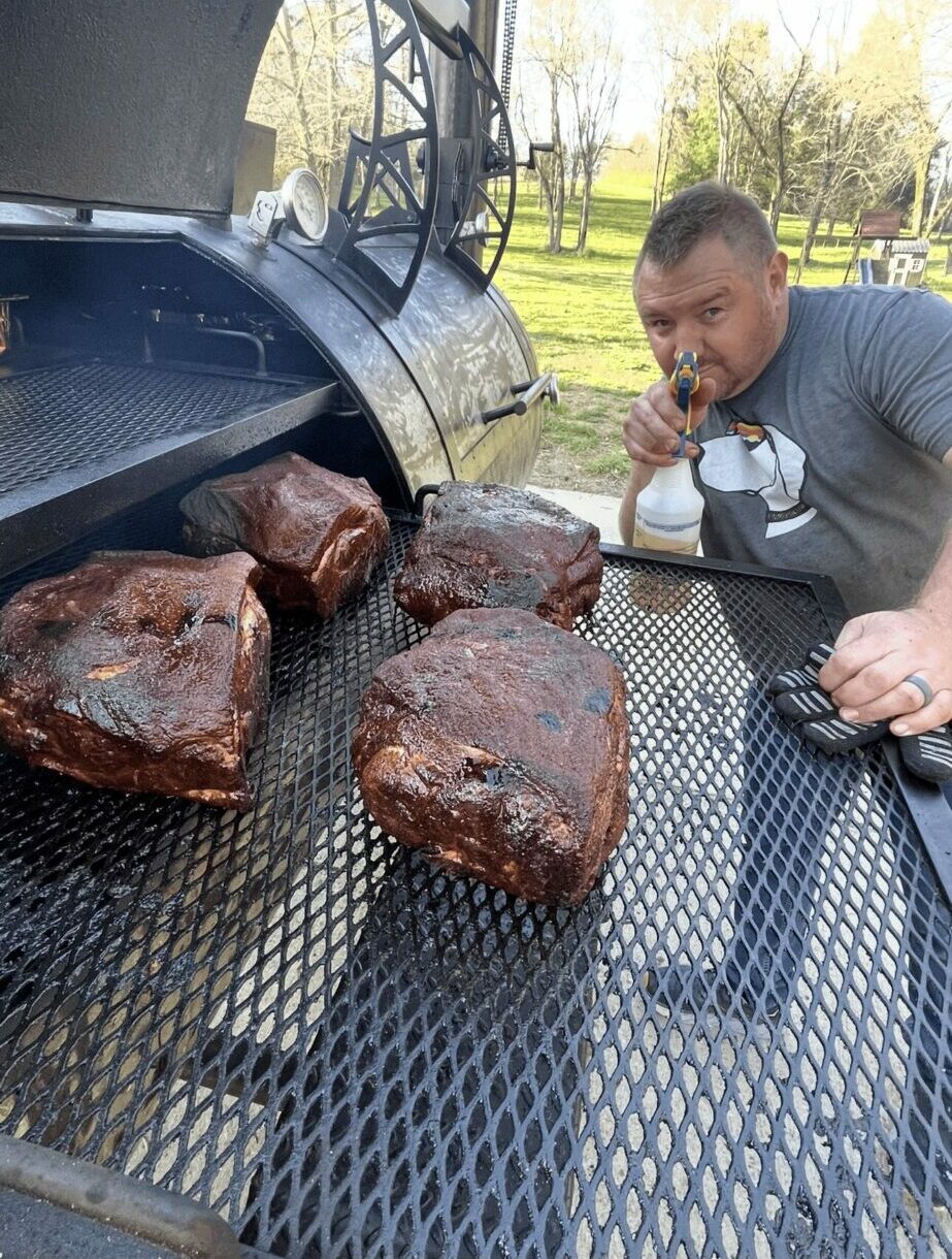 Man leaning on a table with large smoked meat pieces outdoors, trees and grass in background, sunny day.