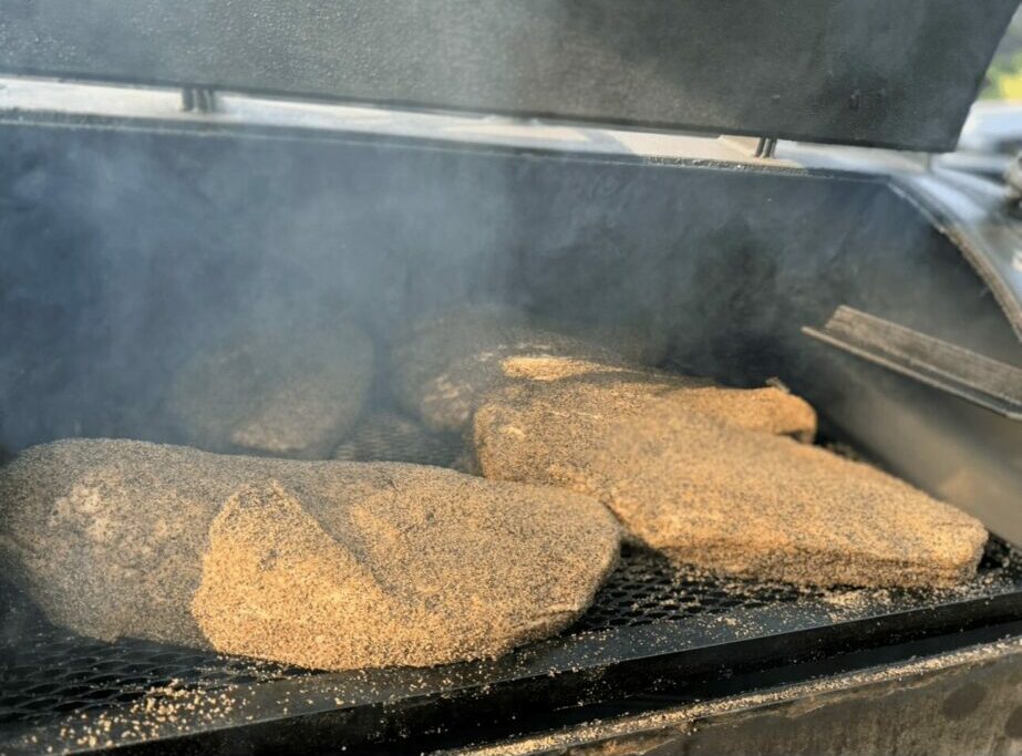 Three pieces of meat cooking on a grill with smoke rising, outdoors, with a metal lid partially open.