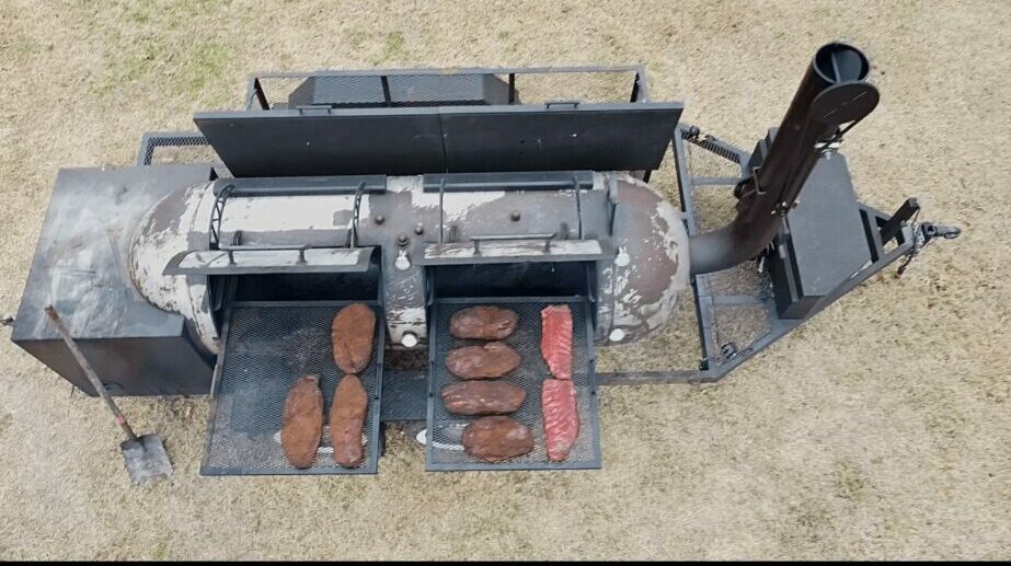 Top-down view of a machine with a black barrel, metal components, and bricks on a sandy surface.