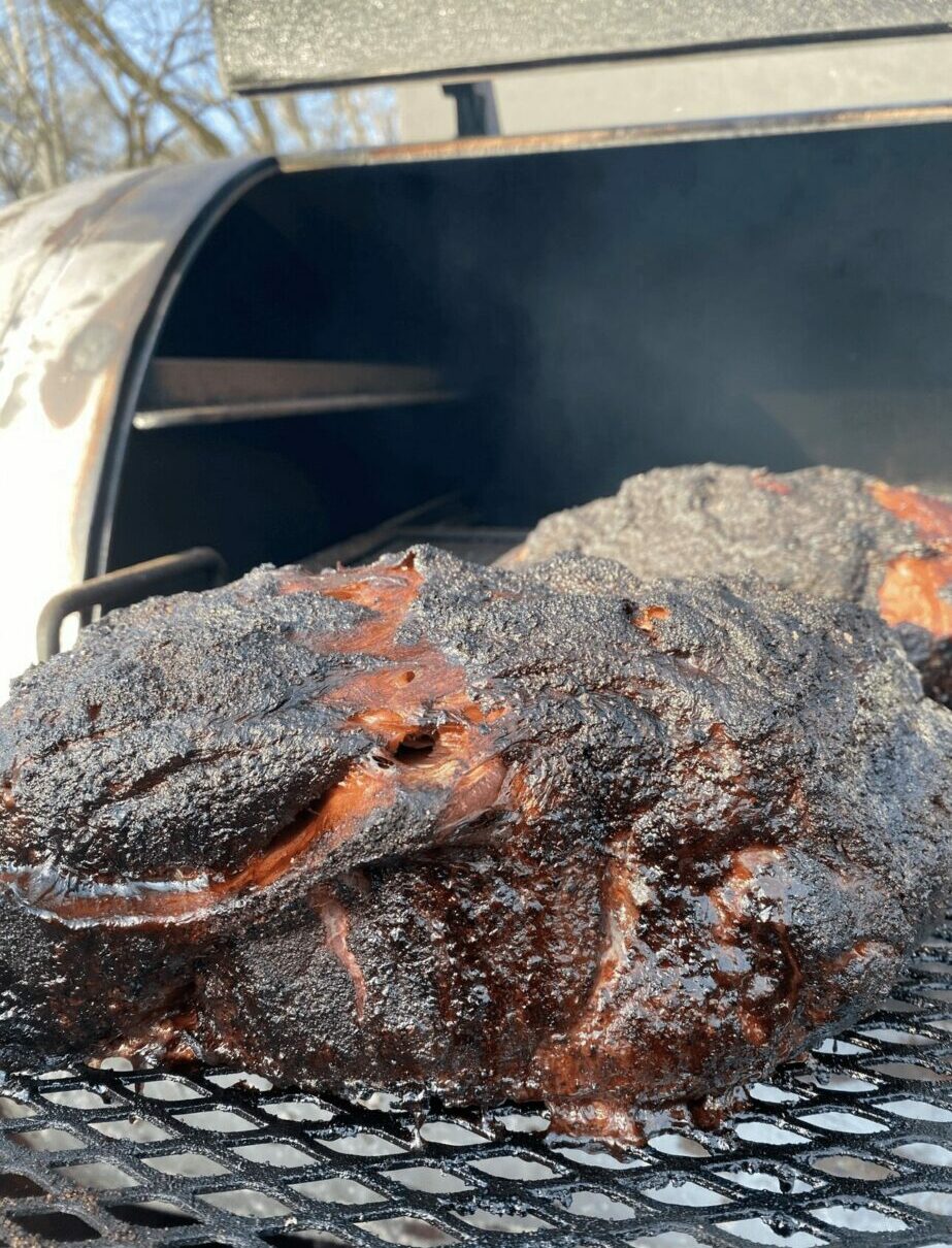 Smoked or grilled large piece of meat with a dark, charred exterior on a grill, outdoors with a blue sky.