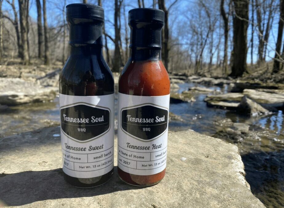 Two bottles of hot sauce on a rock near a stream with trees in the background.